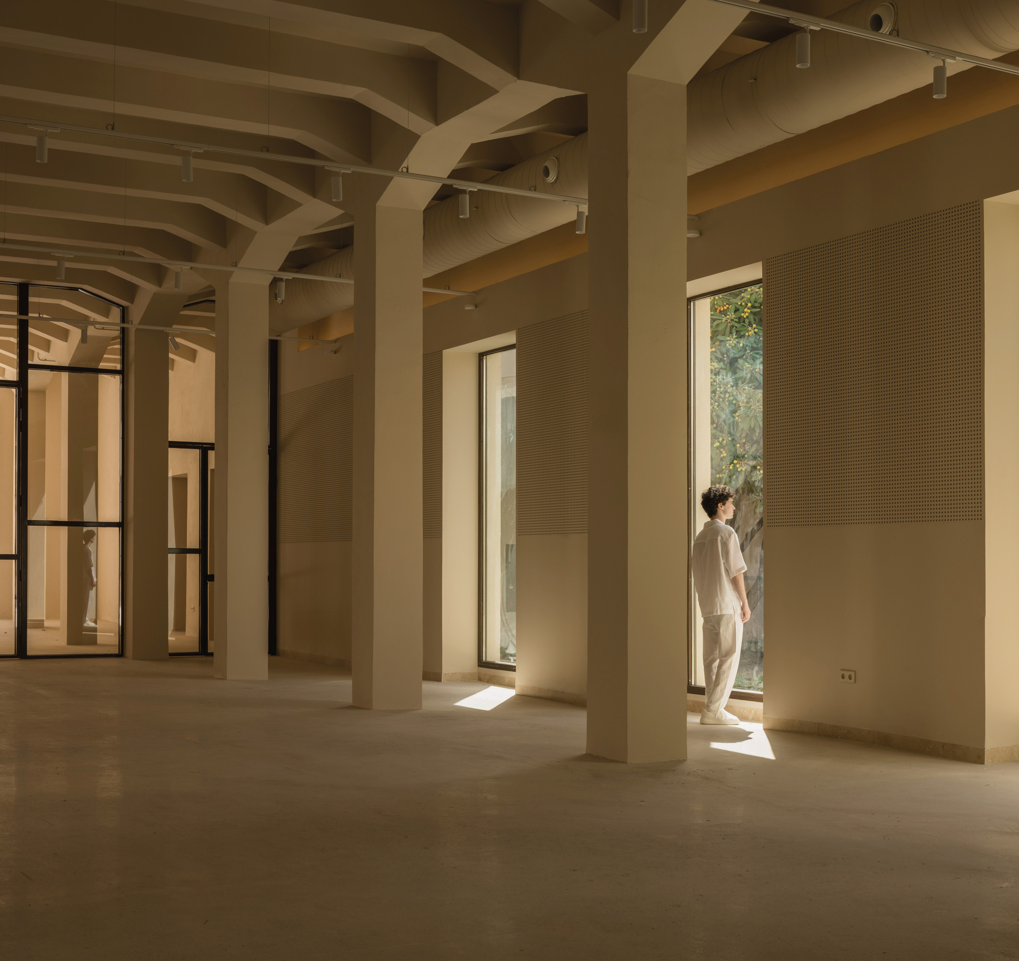 Gallery space with pale arched ceiling beams and a figure framed in a doorway opening to greenery
