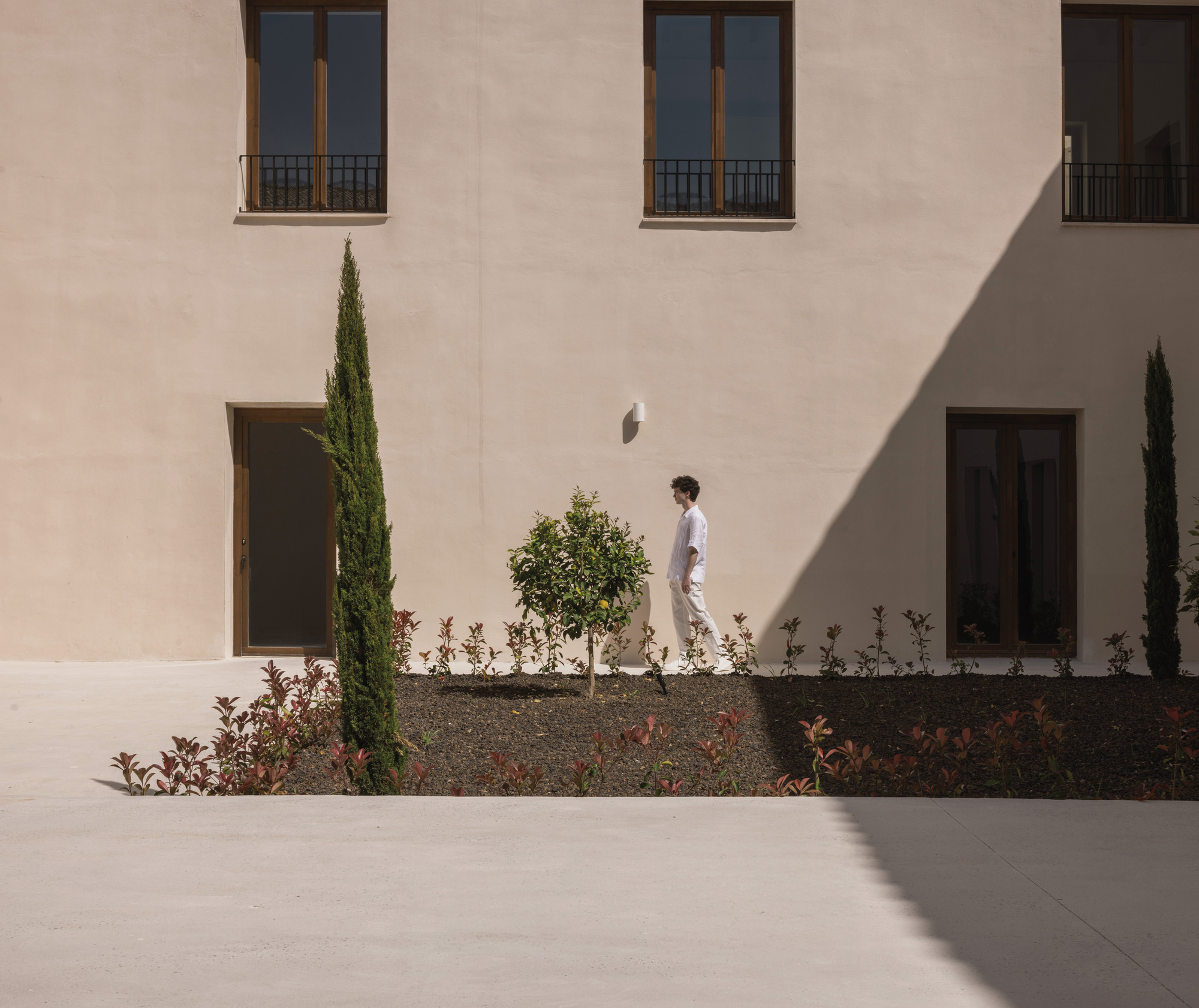 Courtyard view showing a person walking past planted beds with cypress and stucco facades in shadow