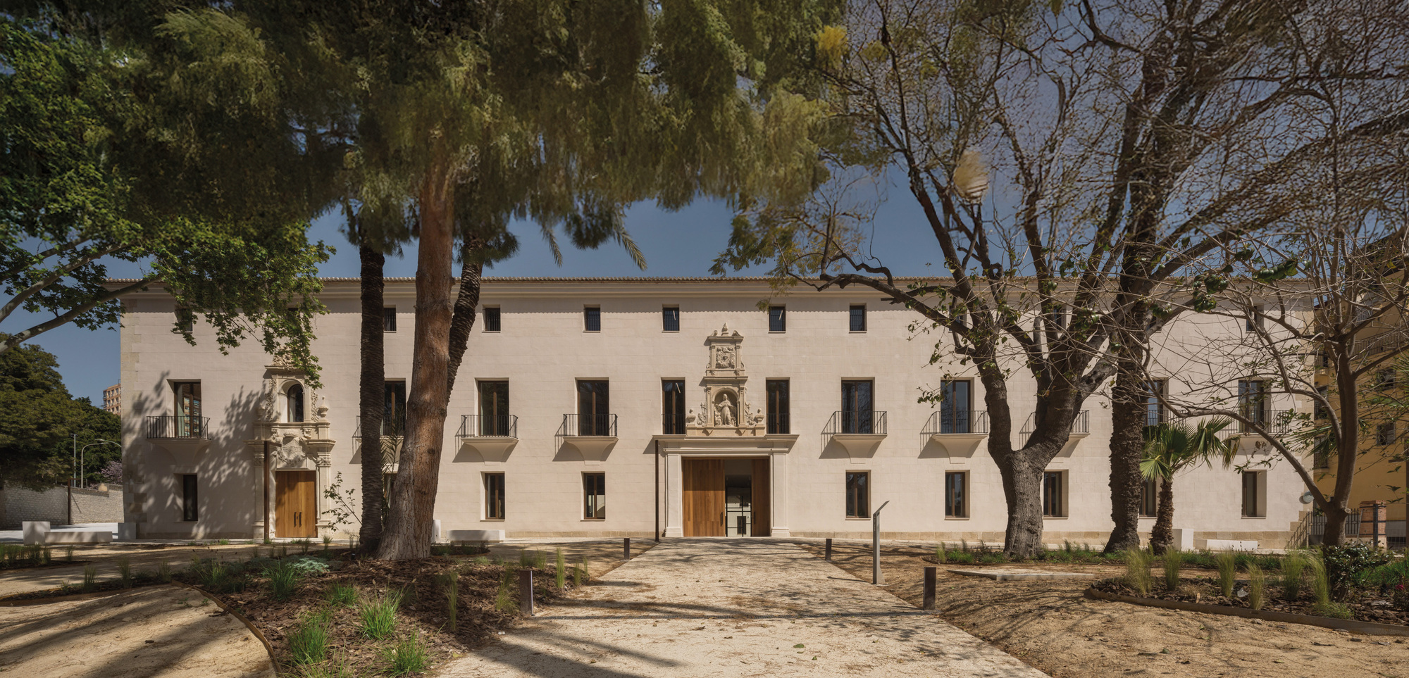 Street facade with ornamental stone portal framed by mature trees casting shadows on gravel forecourt