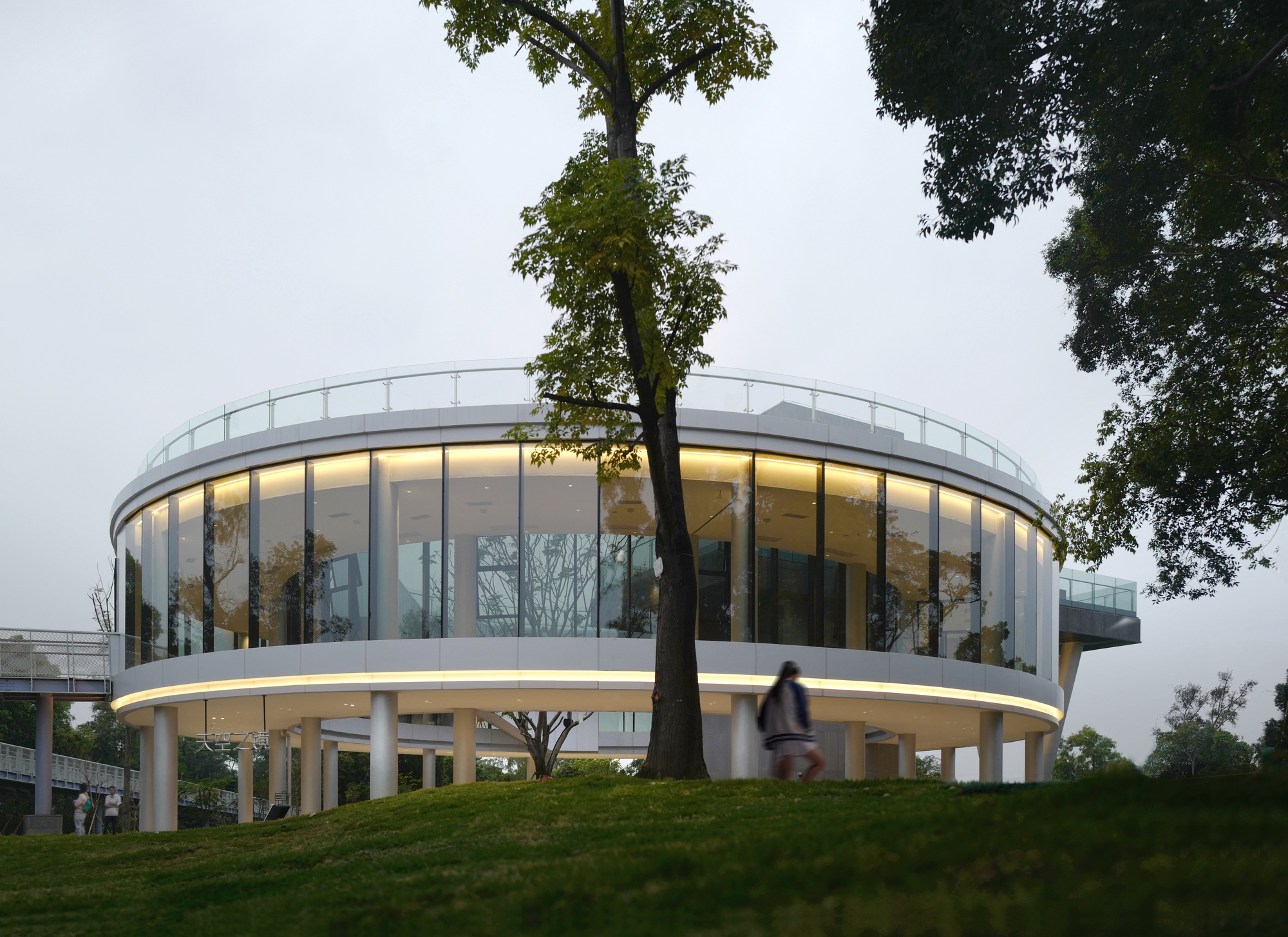 Curved glass facade of the cylindrical building at dusk with interior lighting visible and a cyclist passing by