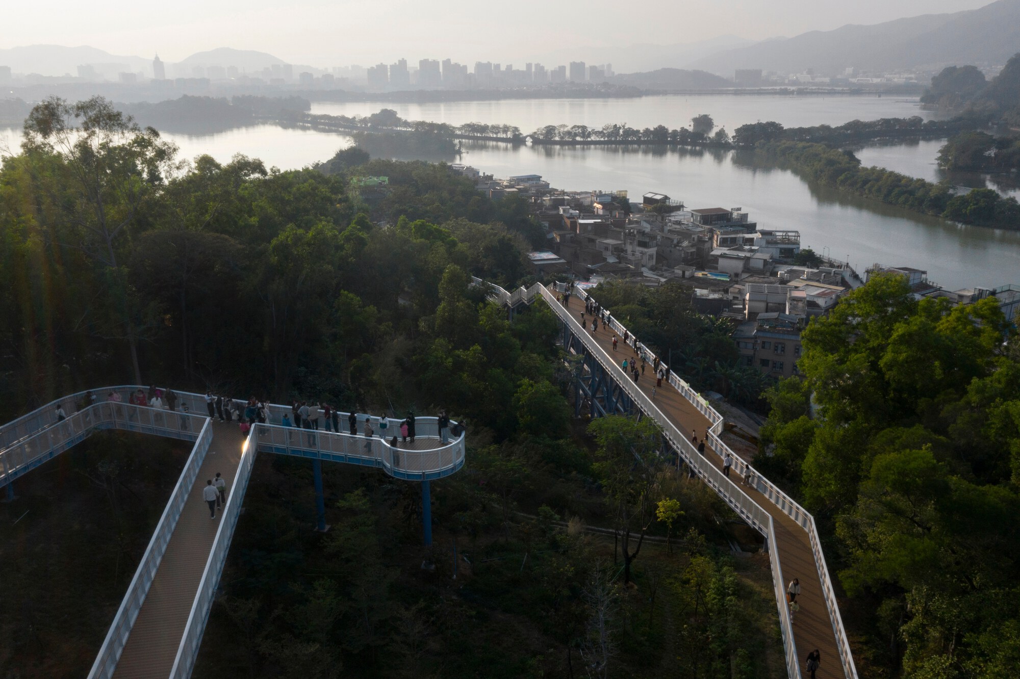 Elevated walkways zigzagging through forested parkland toward the river and distant mountains in early morning light