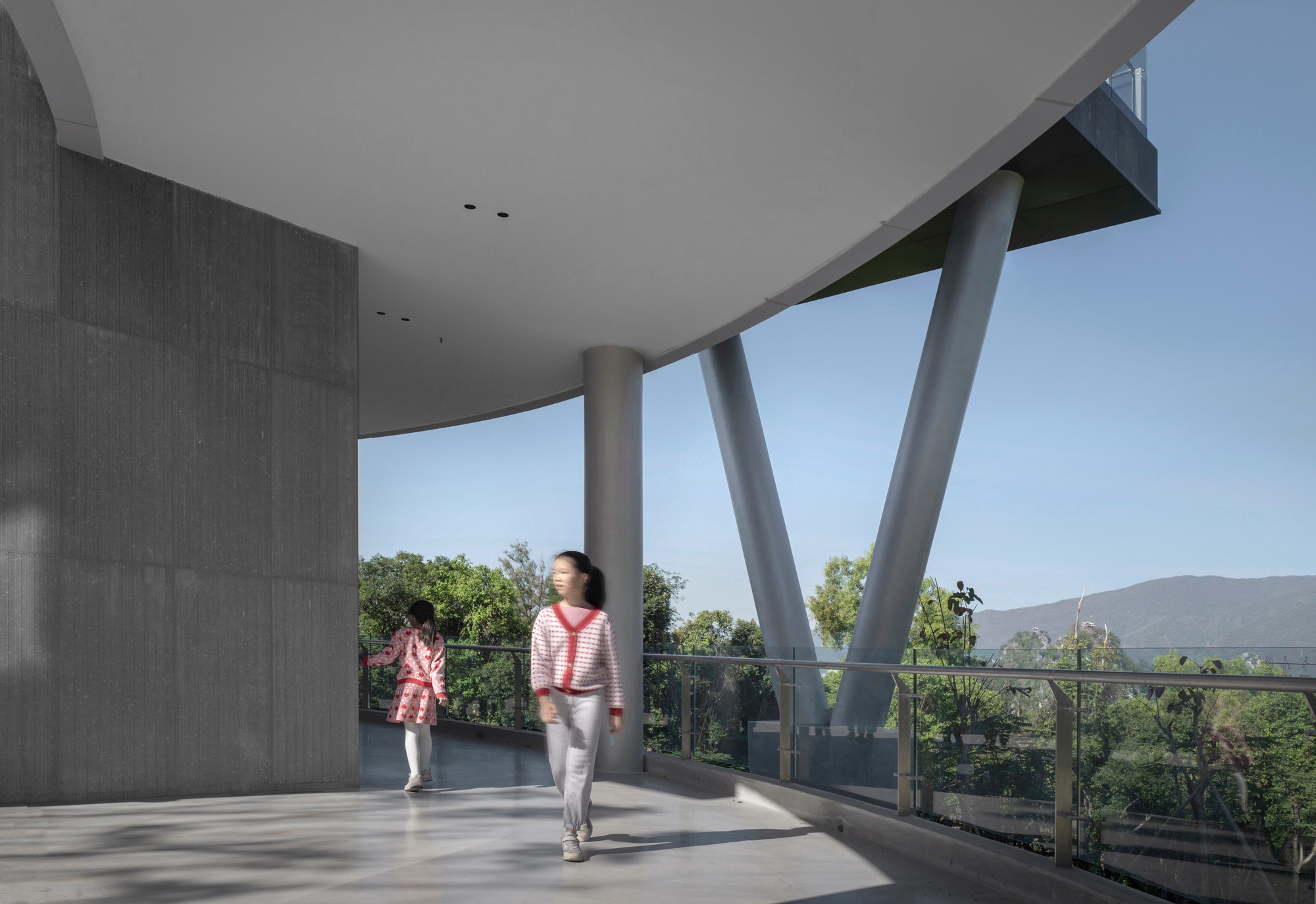 Covered terrace with angled concrete columns and glass railing as two figures walk across the polished floor