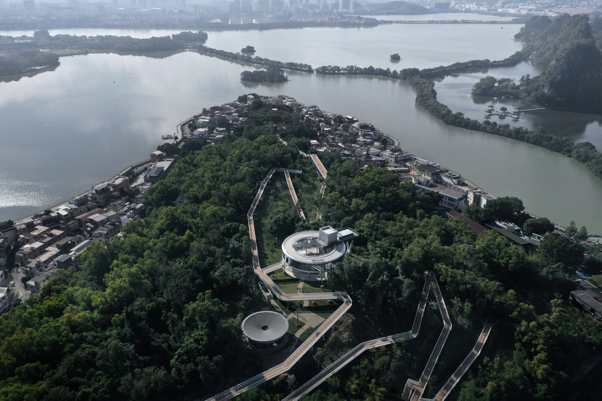 Aerial view of circular pavilions and pathways threading through forested island surrounded by lake water