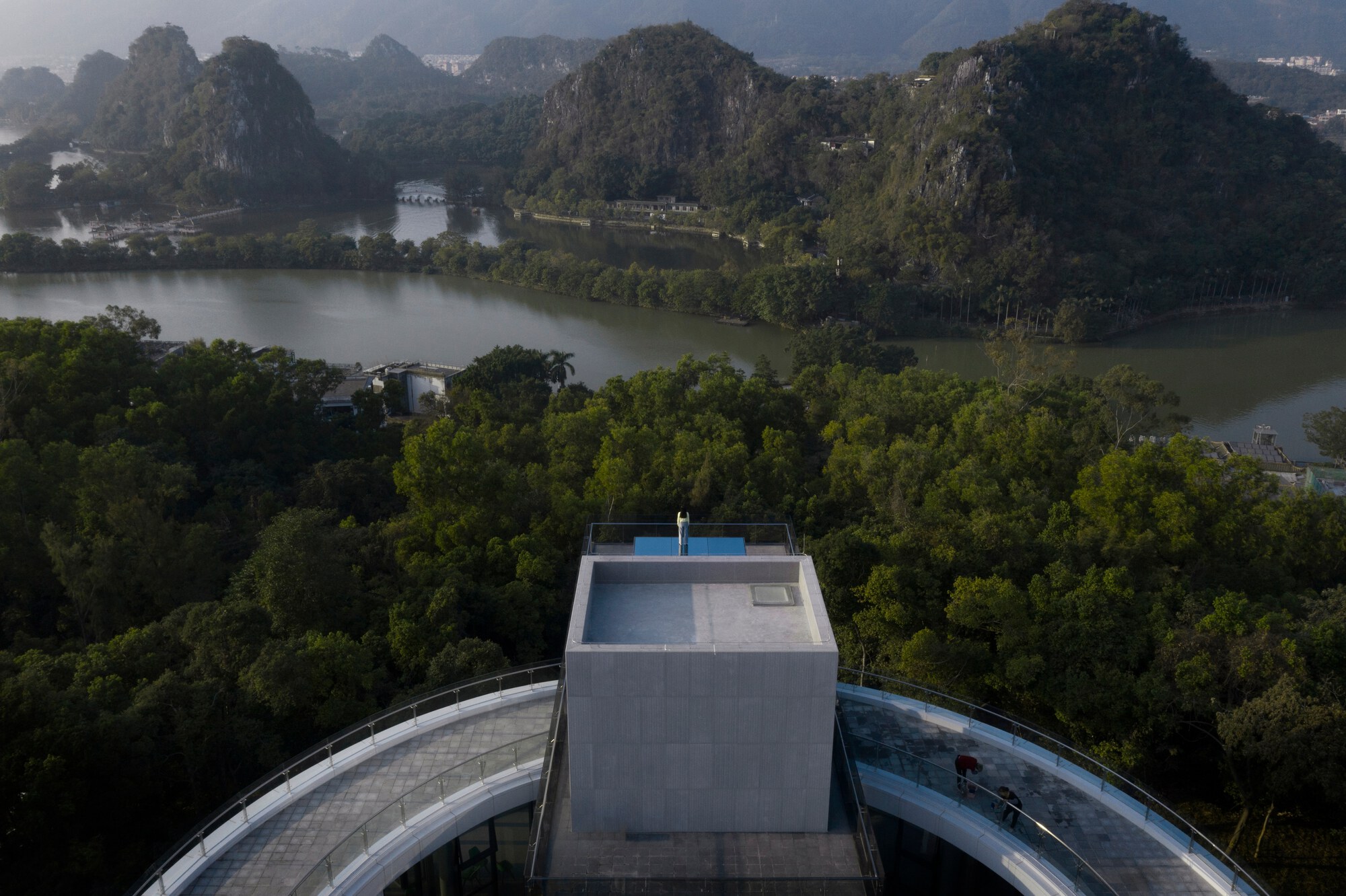 Rooftop pool terrace atop concrete volume overlooking forested valley with karst mountains and lake in morning mist