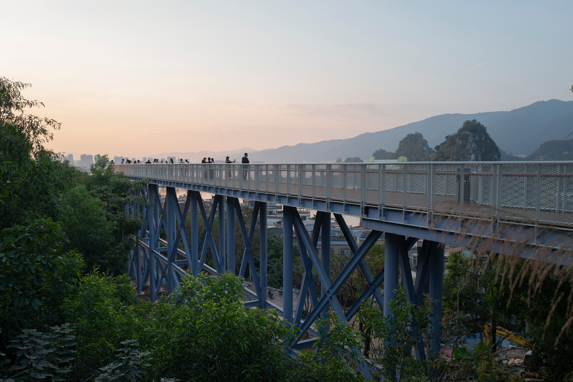 Steel pedestrian bridge with diagonal truss supports crossing over dense greenery at sunset with mountains beyond