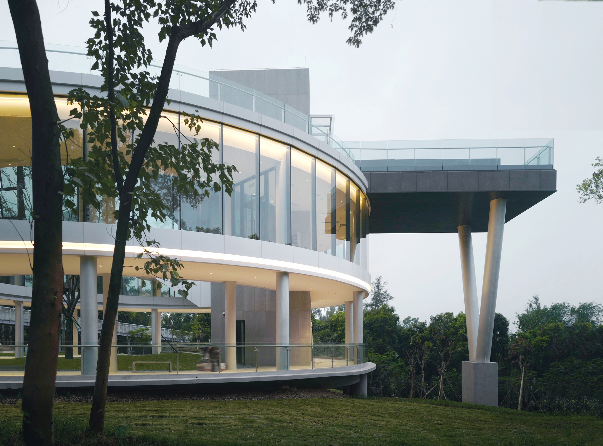 Curved glass pavilion with white columns on a grassy slope framed by deciduous trees