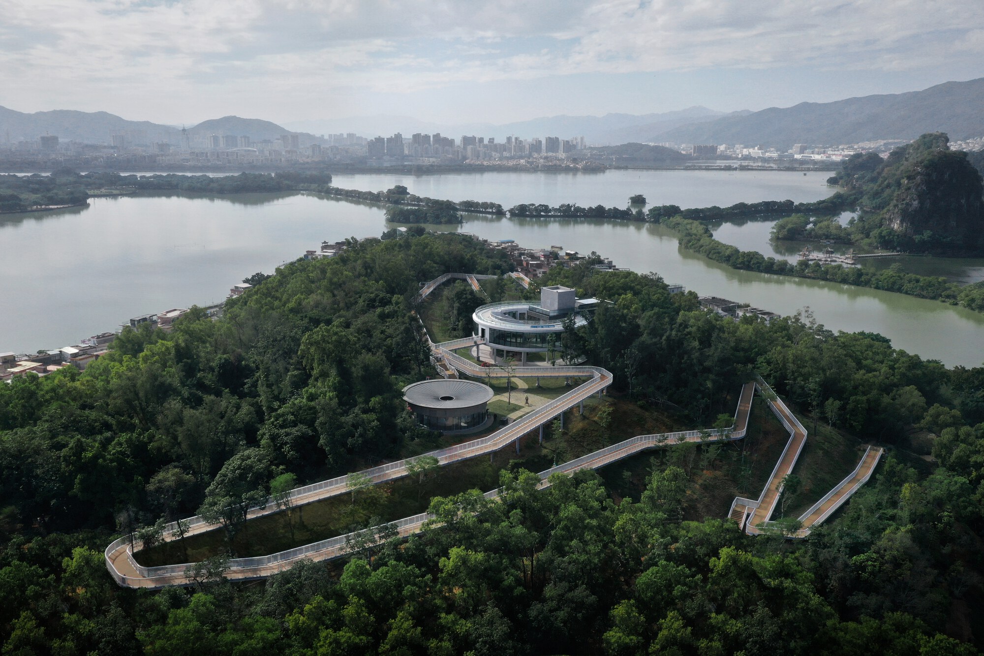 Aerial view of a zigzagging walkway connecting a circular pavilion on a forested peninsula surrounded by water