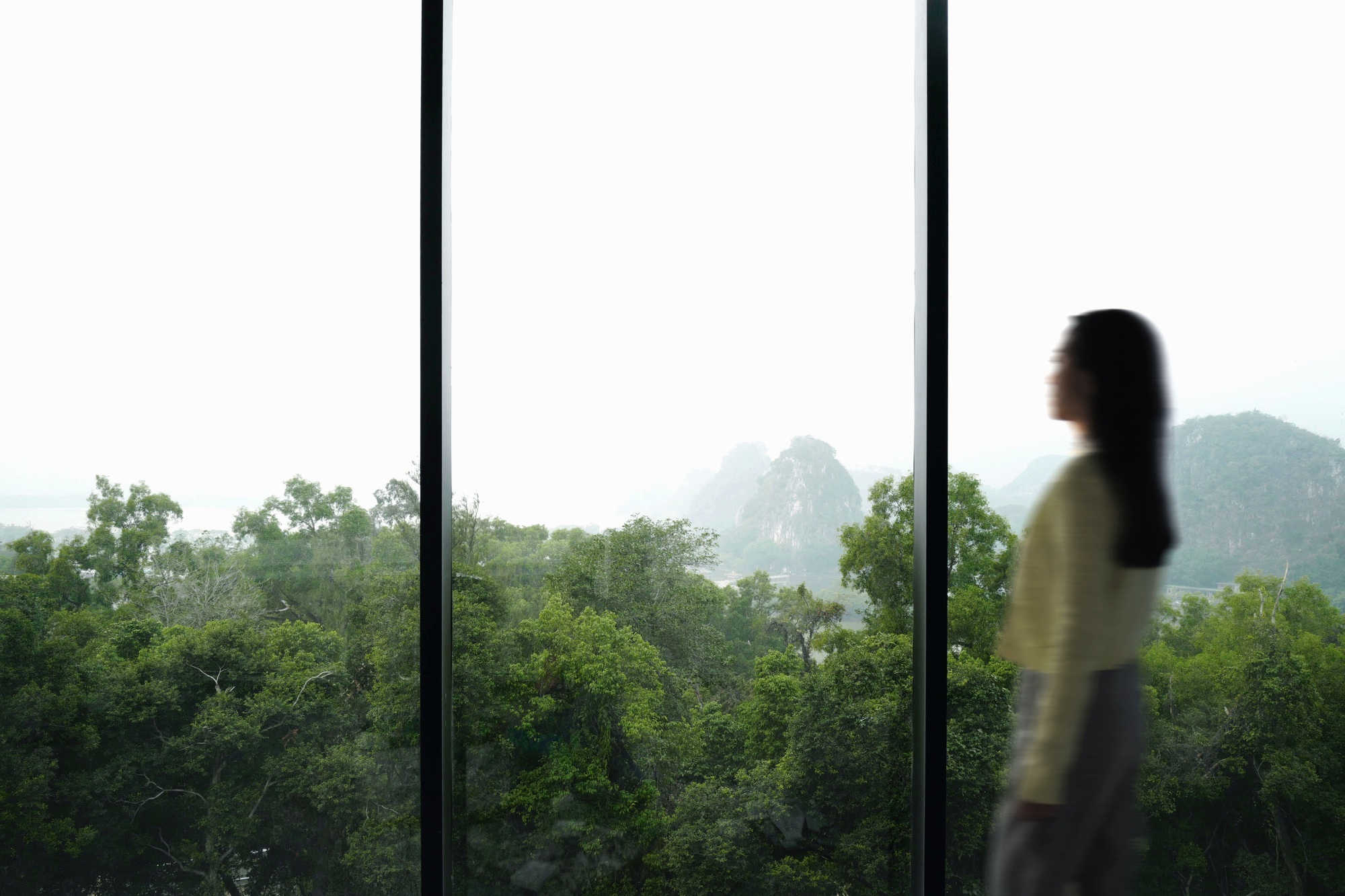 Floor-to-ceiling window framing a forested valley with karst peaks visible through morning haze as a figure passes