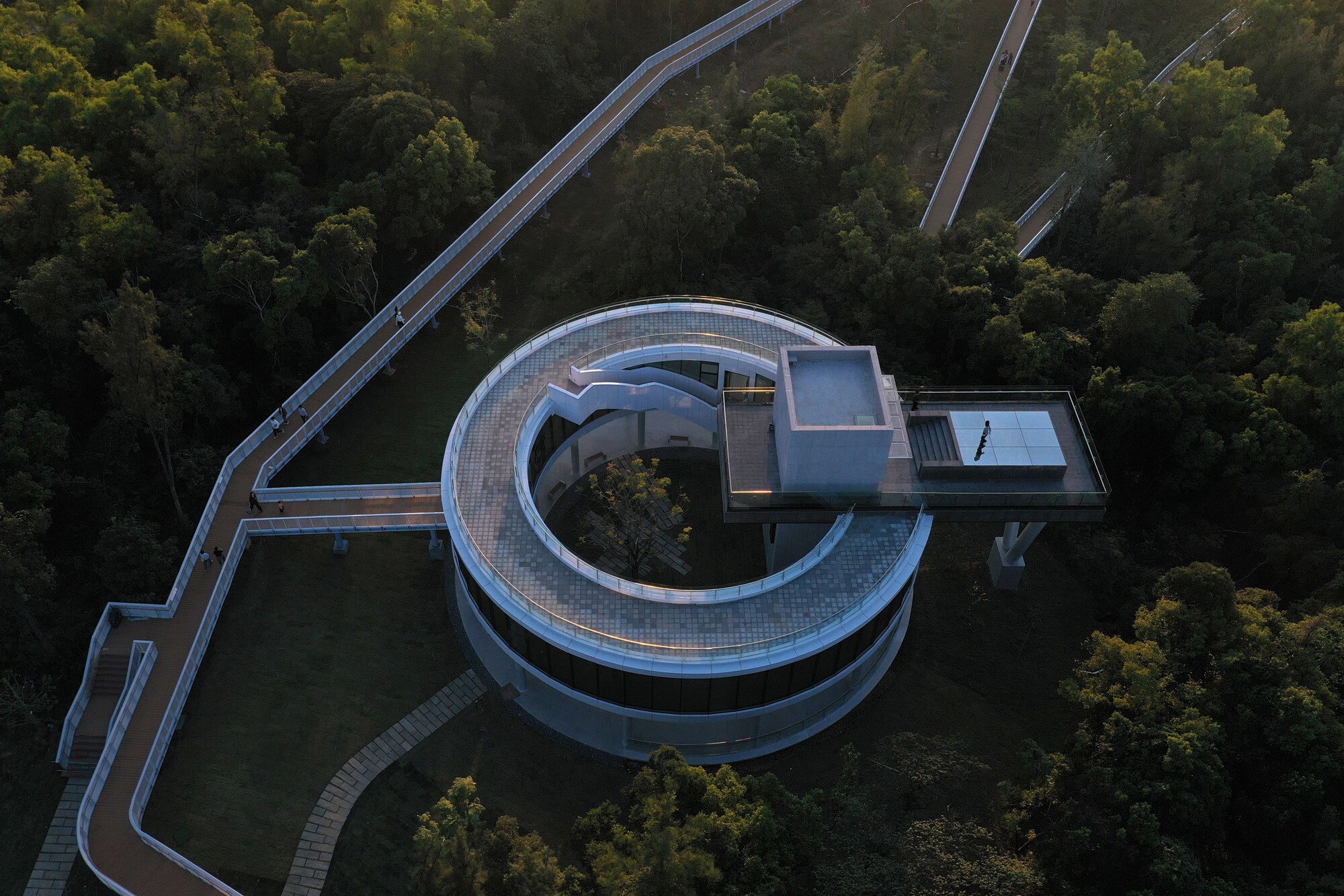 Drone view of the spiral building with curved walkways and bridges extending into the surrounding wooded hillside