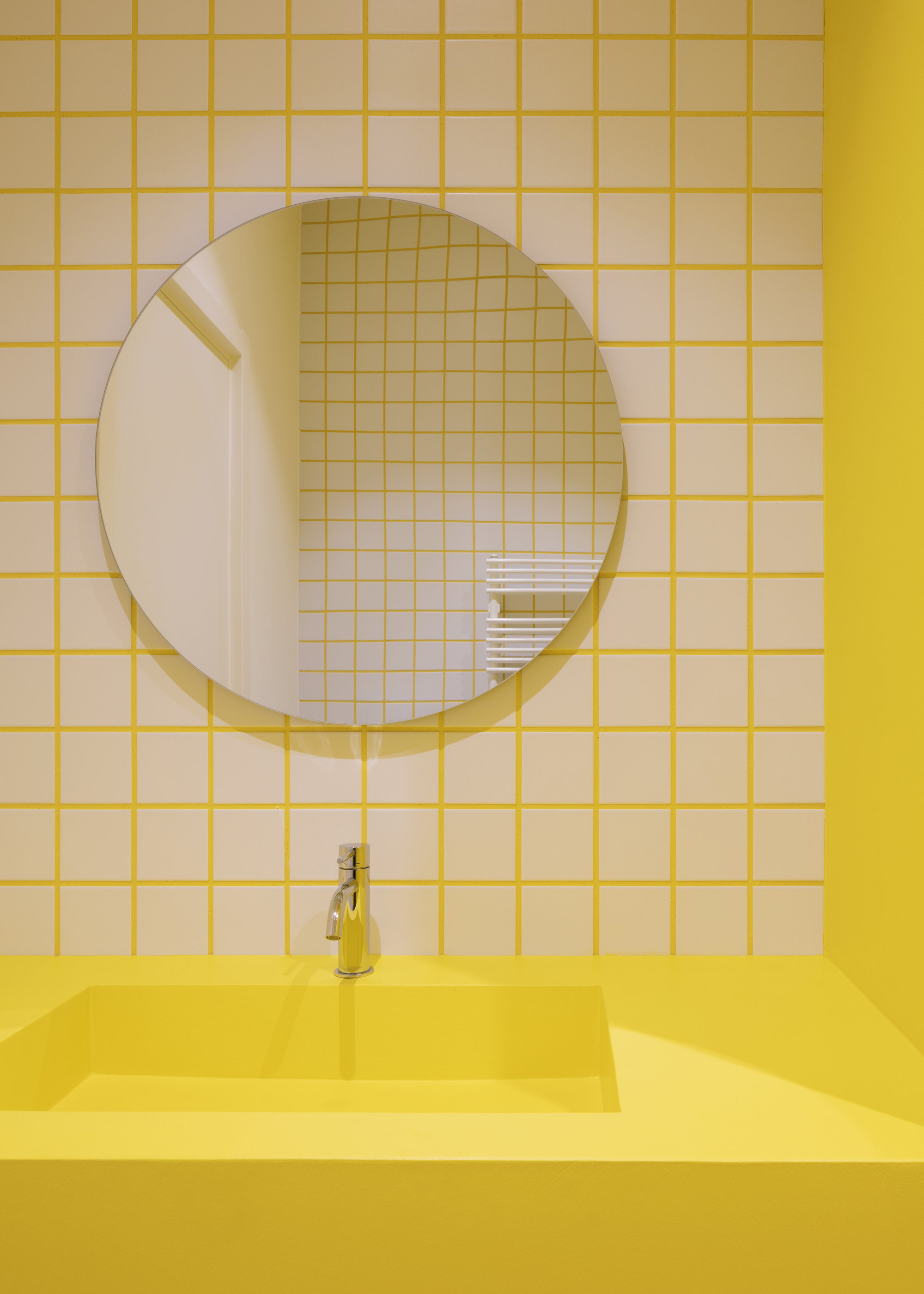 Yellow square-tiled bathroom wall with round mirror above yellow integrated sink and counter