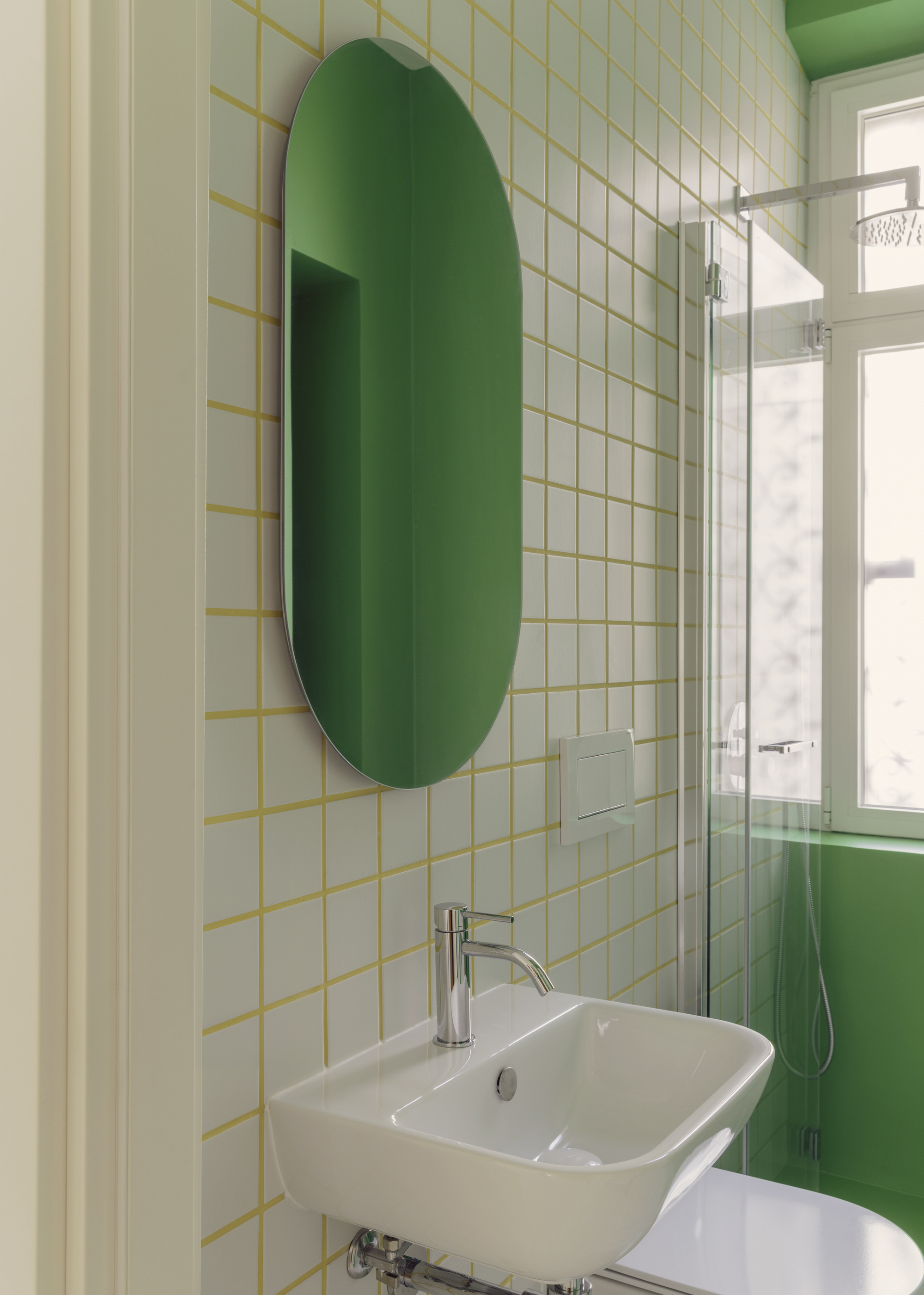 Green oval mirror on white square-tiled wall with yellow grout beside a window