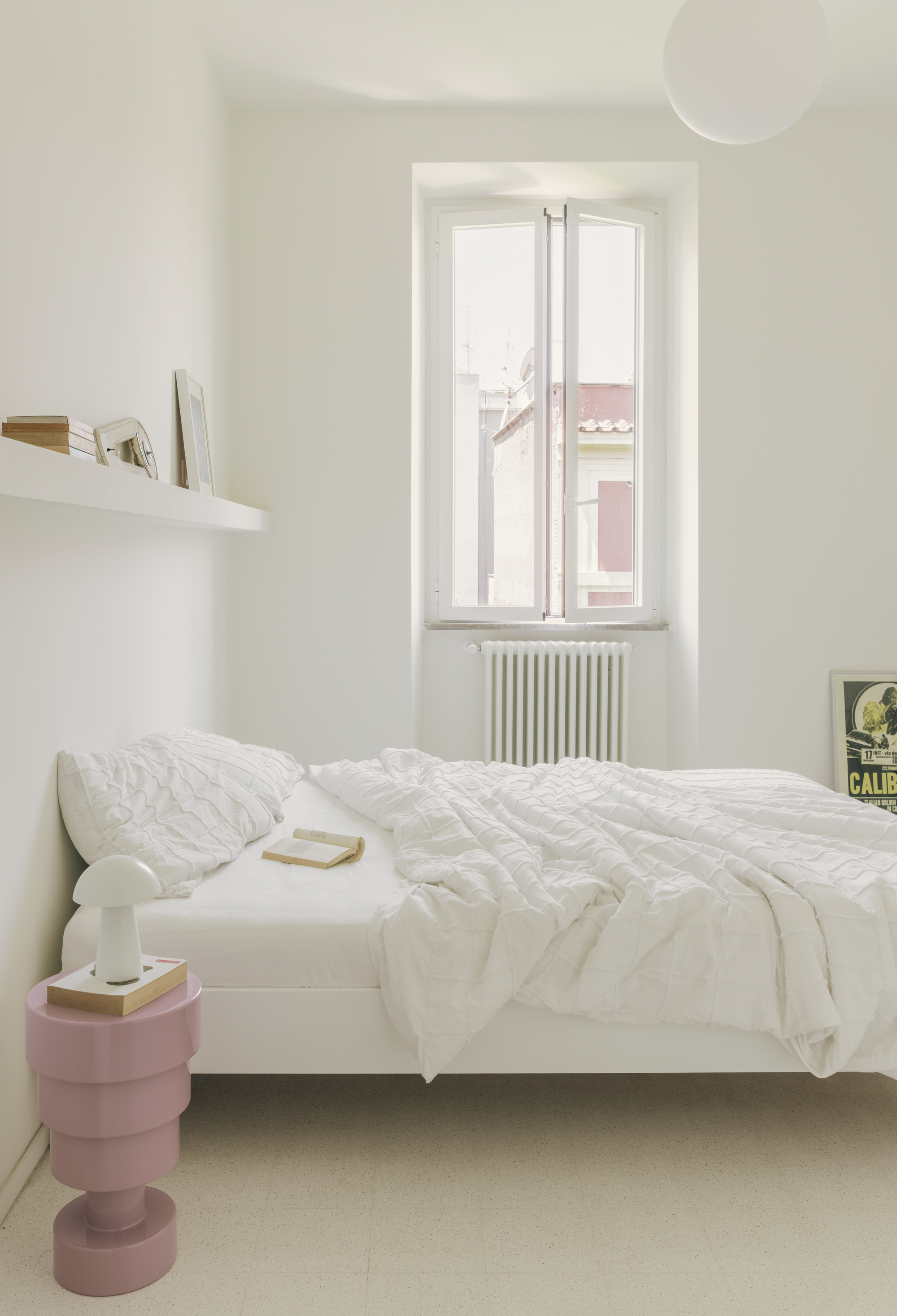 Bedroom with arched window revealing rooftops, white radiator below and pink cylindrical bedside table