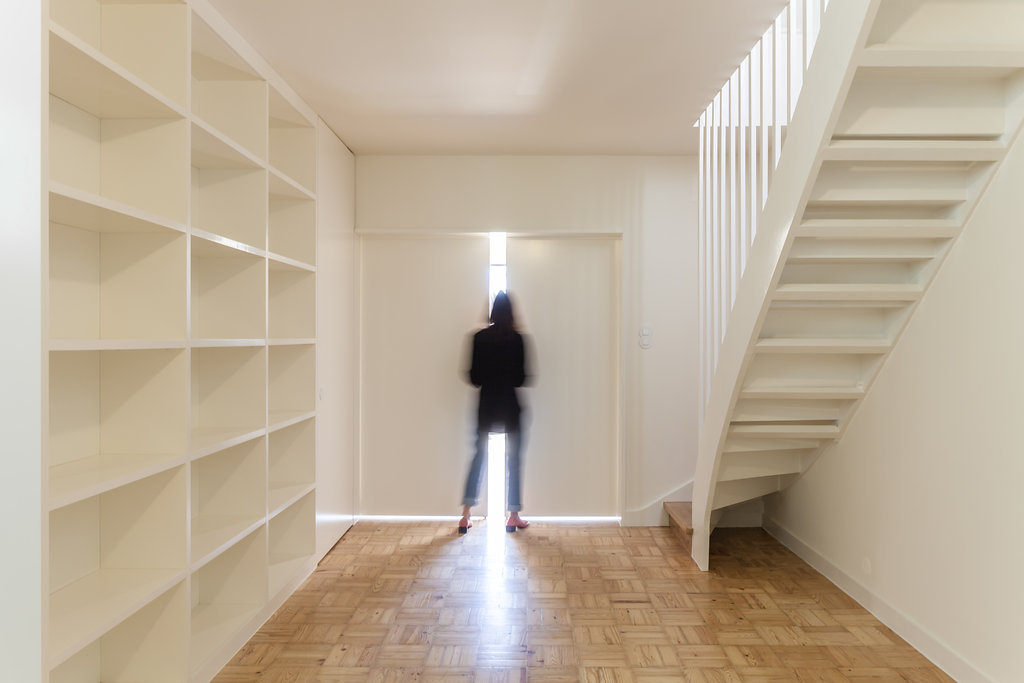 Open white shelving beside a white open-riser staircase, a blurred figure standing at a sliding door with light slipping through the gap