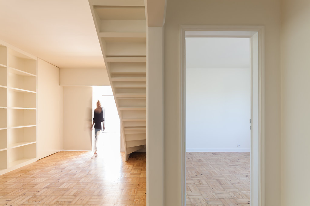 Underside of the white staircase beside built-in shelves, a blurred figure walking toward bright light over parquet floors and an open doorway