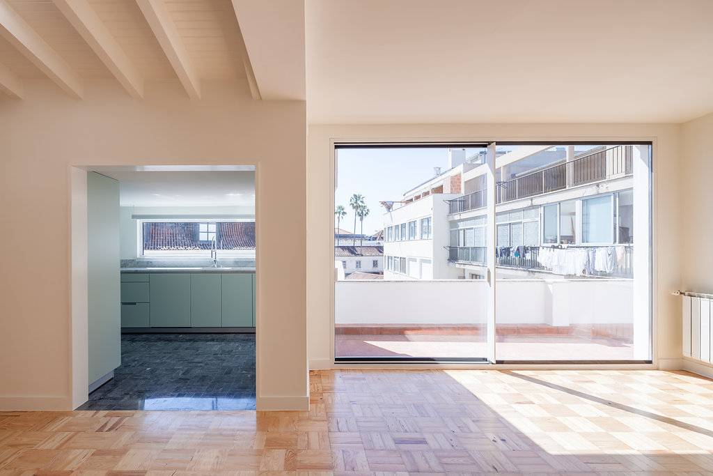 Living room parquet in sunlight with a sliding glass door to a terrace, and a pale green kitchen visible through an opening