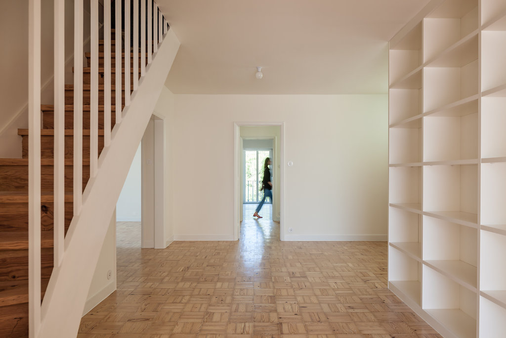 Hallway with a timber stair and white balusters, tall built-in shelving, and a blurred figure crossing a distant doorway over parquet