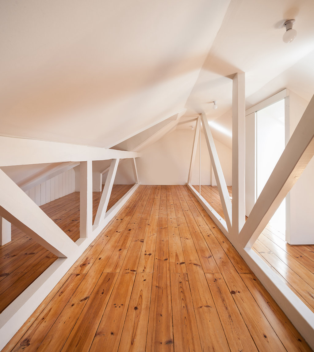 Attic loft with white timber trusses and pine plank flooring lit by soft daylight from a side opening