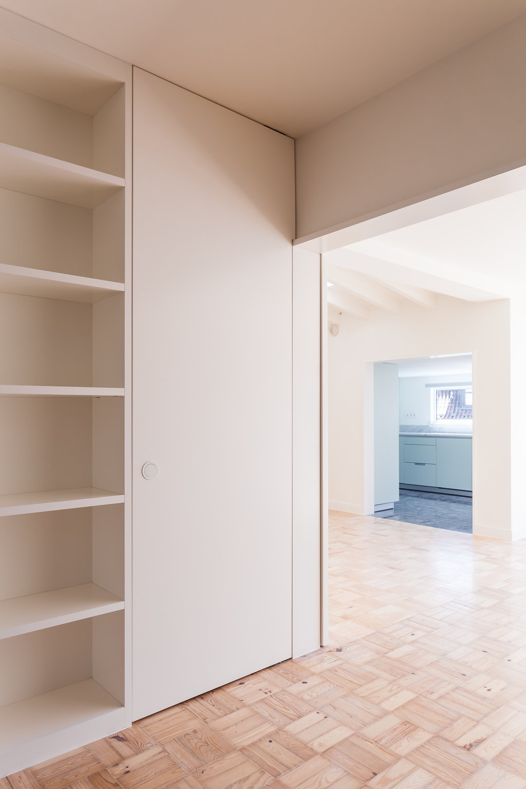 Built-in white shelving and flush sliding door beside a doorway opening onto a kitchen, over parquet flooring