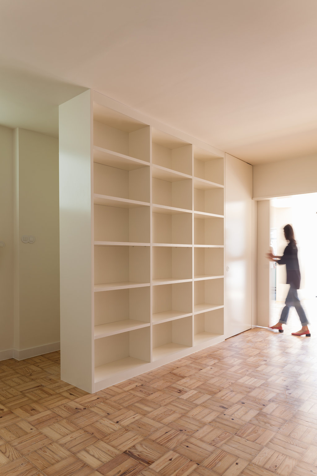 Floor-to-ceiling white bookcase dividing a parquet-floored room, with a blurred person walking through a sunlit doorway