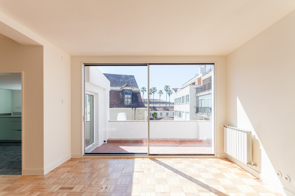 Sliding glass door opening onto a roof terrace with palms and tiled rooftops, sunlight cutting across the parquet floor