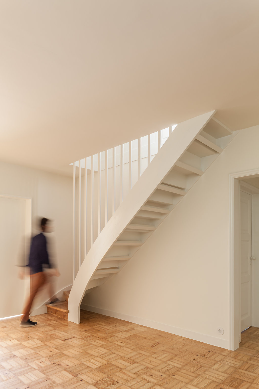 White open-riser staircase with slatted balustrade curving up from a parquet floor, a blurred person walking past