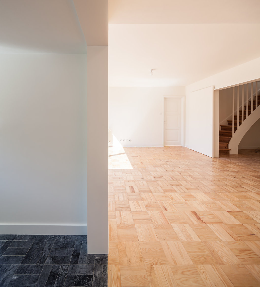 Threshold between dark grey stone tiles and pine parquet, with a curved staircase visible in the sunlit room beyond