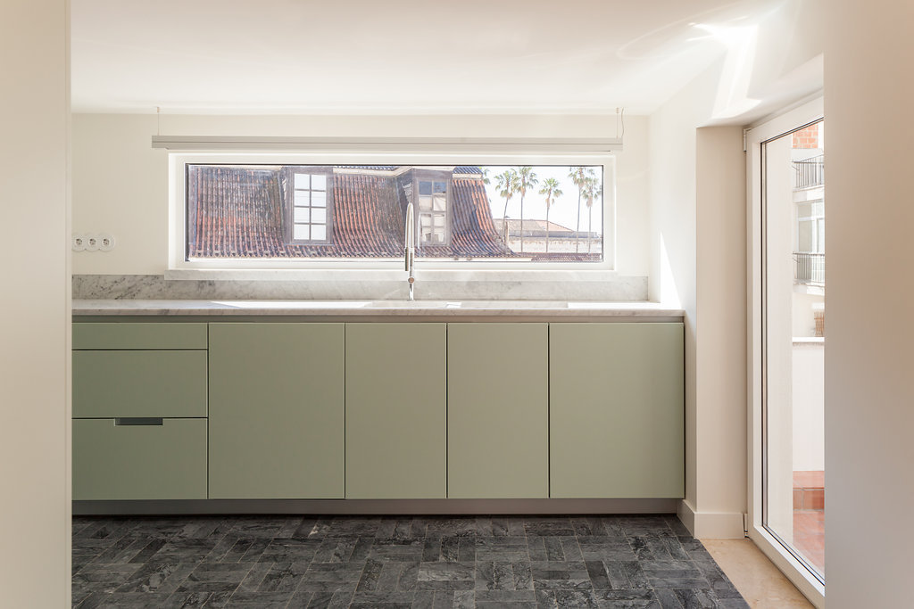 Sage green kitchen cabinets beneath a long strip window framing tiled rooftops and palm trees in midday light