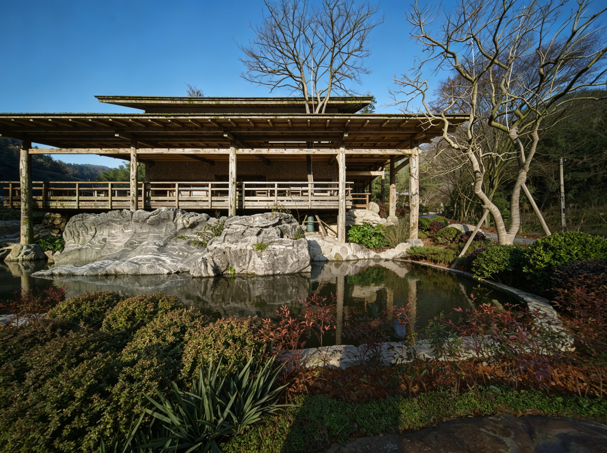 Open timber pavilion with exposed columns and rafters reflected in a pond with large boulders and plantings