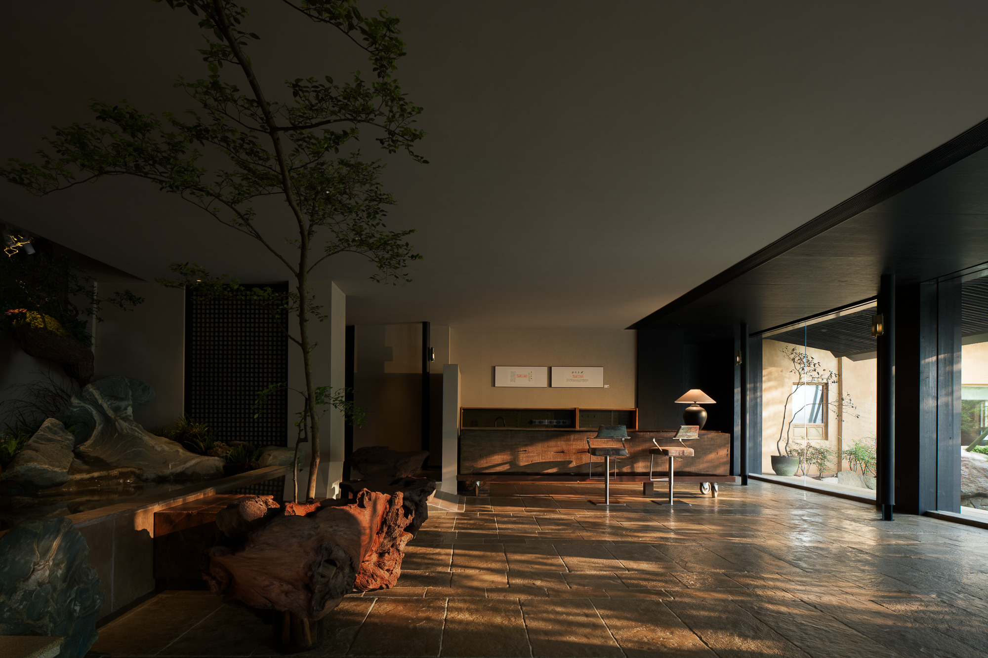 Interior living space with stone flooring and tree planting beneath a light well in afternoon sunlight