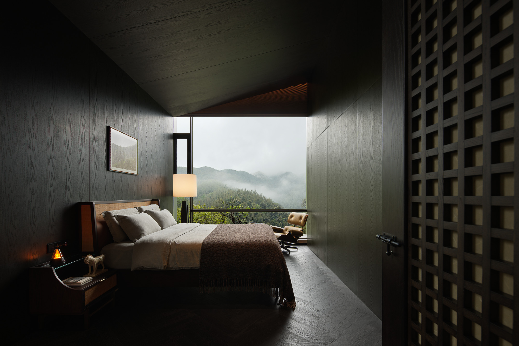 Bedroom with textured dark walls and clerestory window framing mist-shrouded peaks beyond a lounge chair
