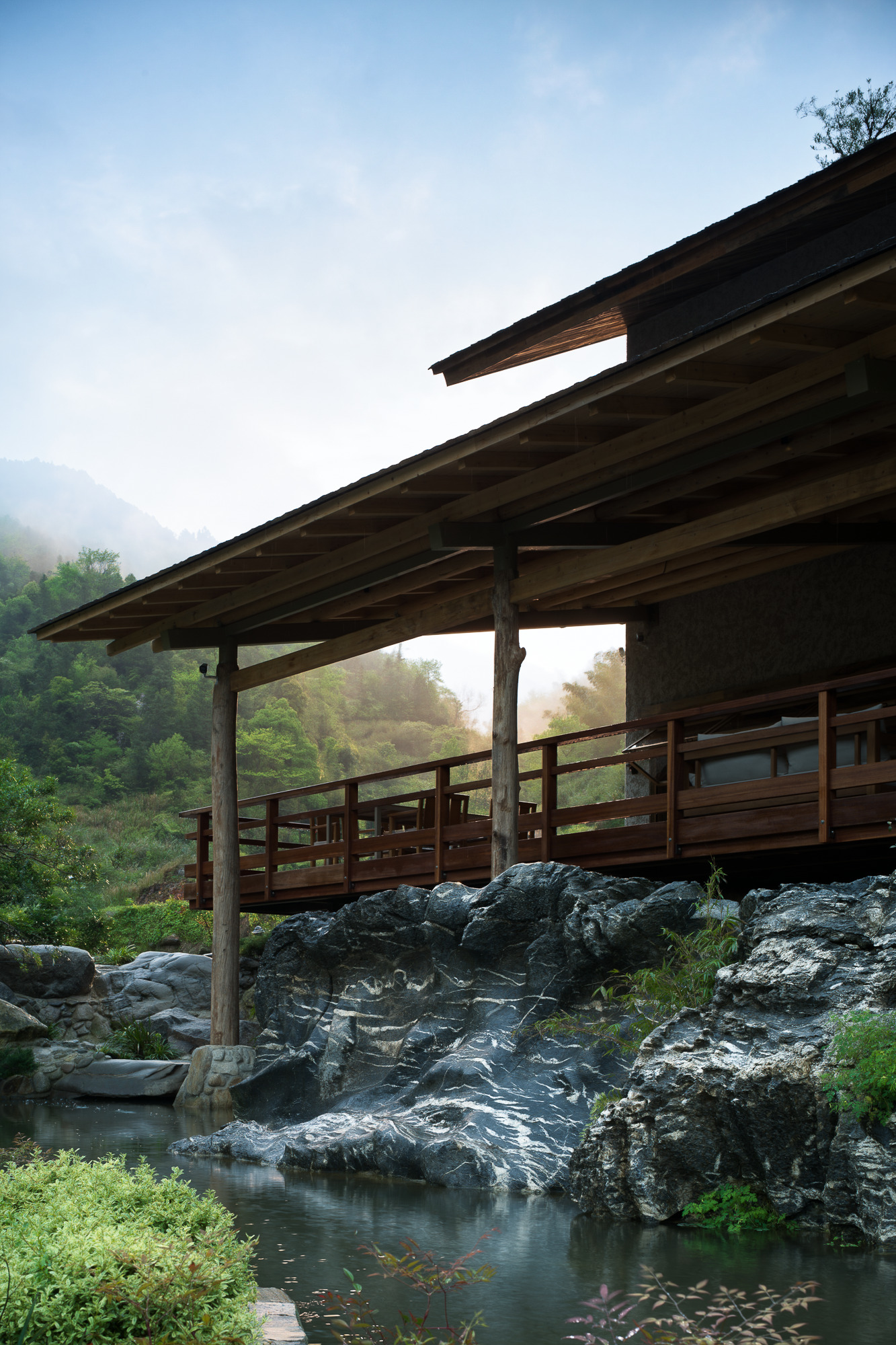 Timber pavilion with exposed rafters spanning a rocky stream in morning mist