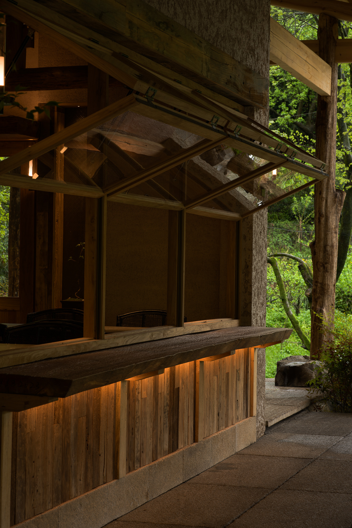 Illuminated timber-clad base beneath glazed facade with exposed tree trunks at dusk