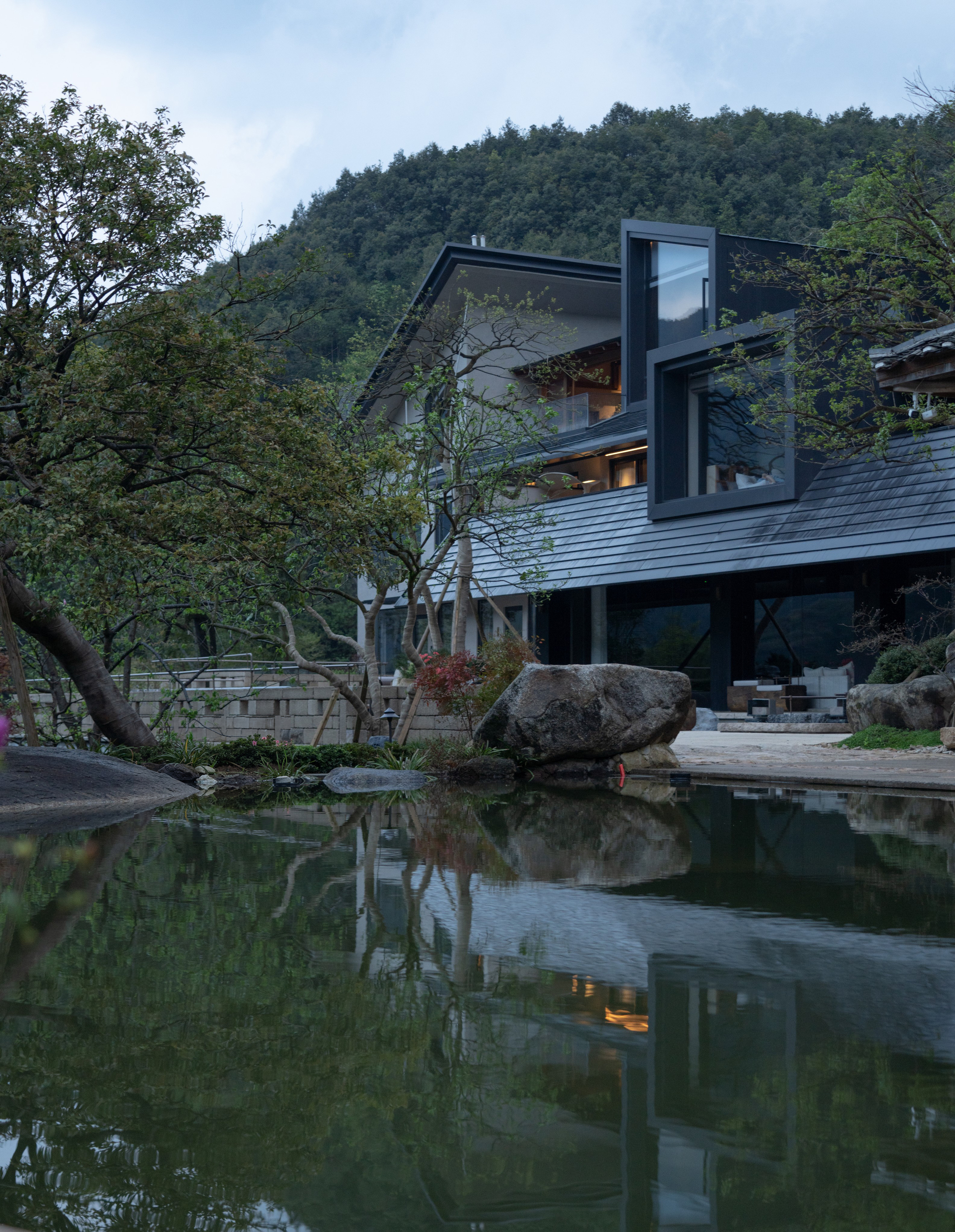 Angled black-framed glazing above pond with boulders and tree reflections at twilight