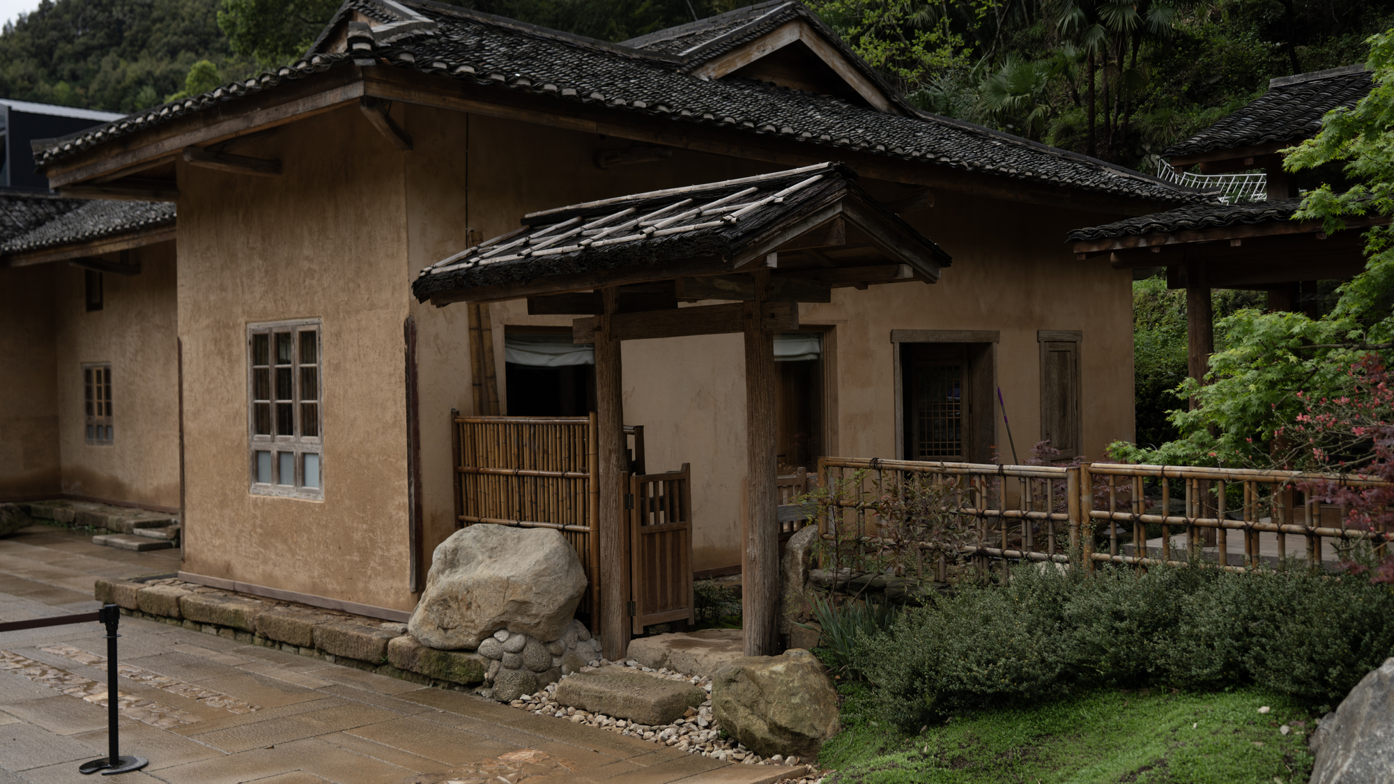 Earthen-walled pavilion with tile roof and bamboo fence beside planted gravel bed