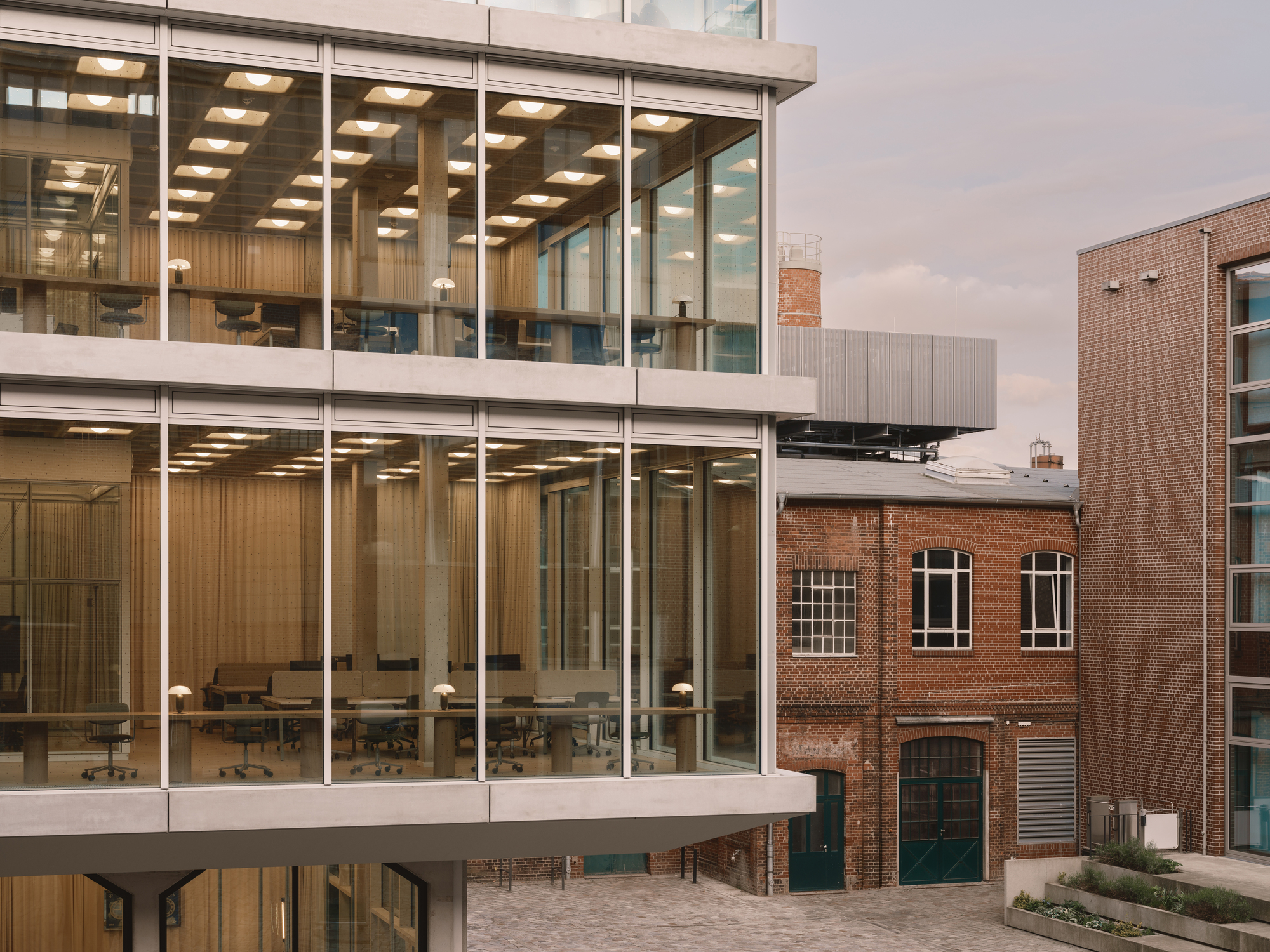Glass-wrapped floors with exposed interior workspaces beside a red brick industrial building under overcast sky