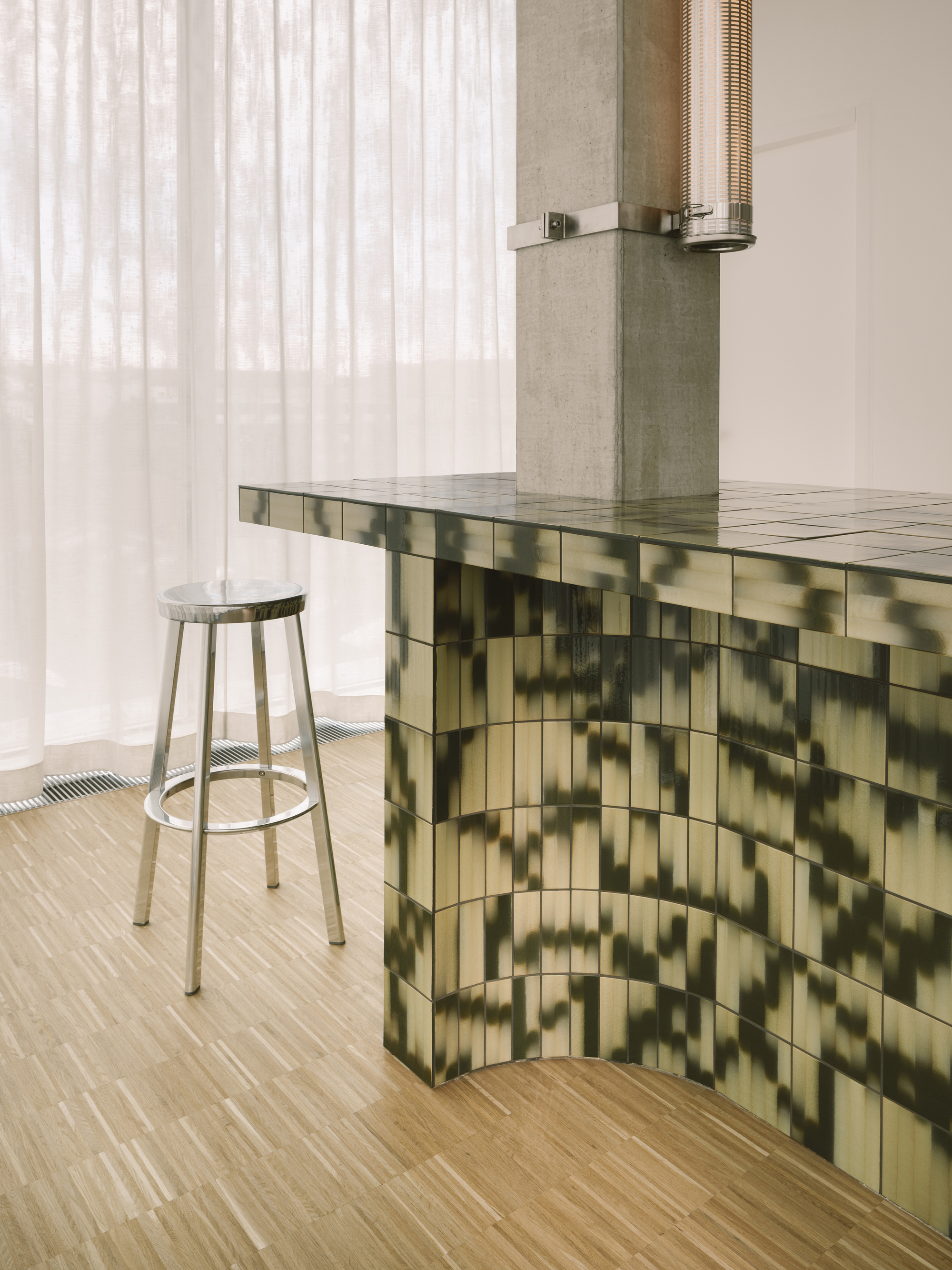 Reception counter clad in green gradient glazed tile with metal stool and sheer curtain backdrop