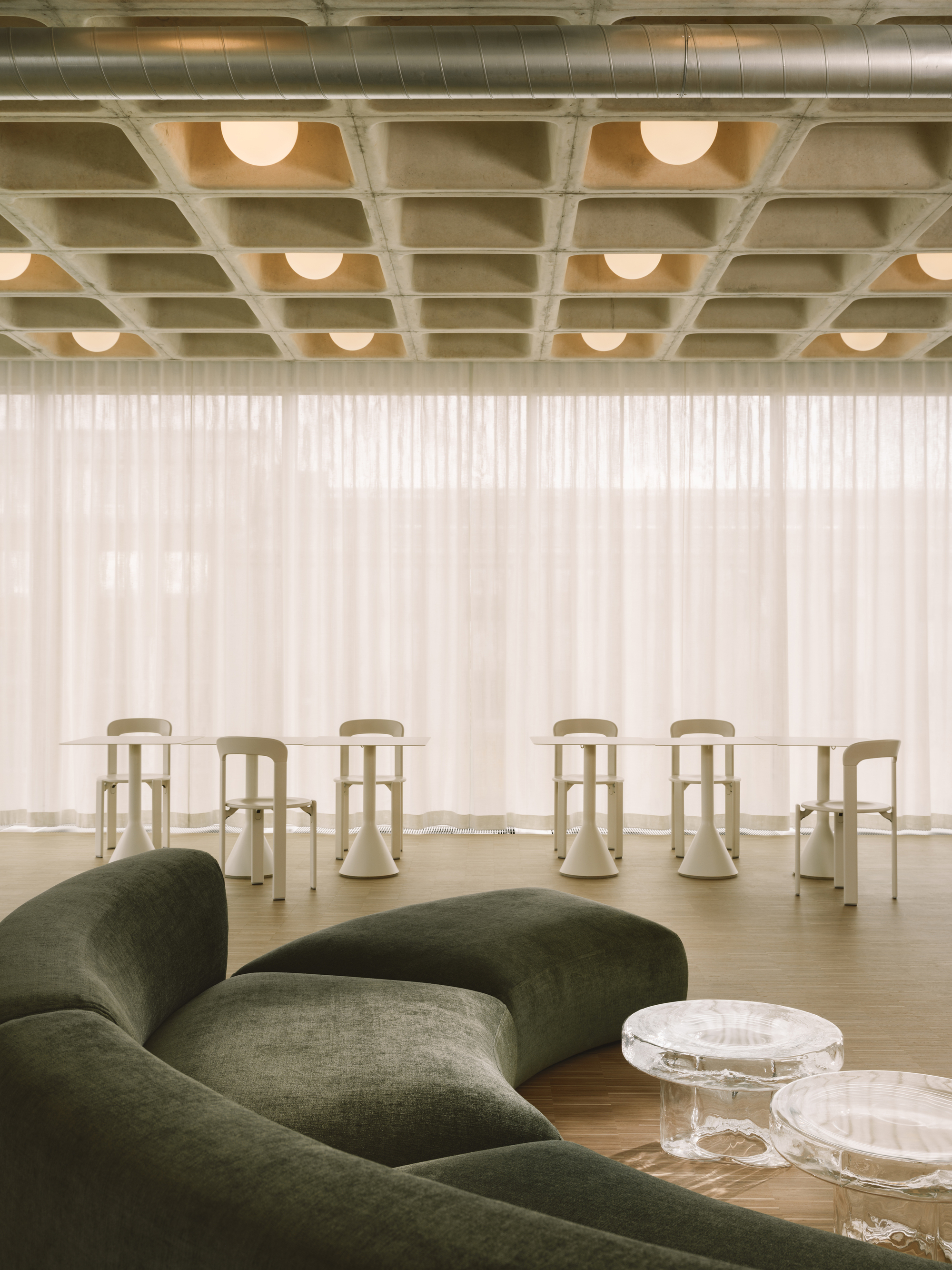 Lounge area with concrete coffered ceiling, pale wood stools and green upholstered seating beneath sheer curtains