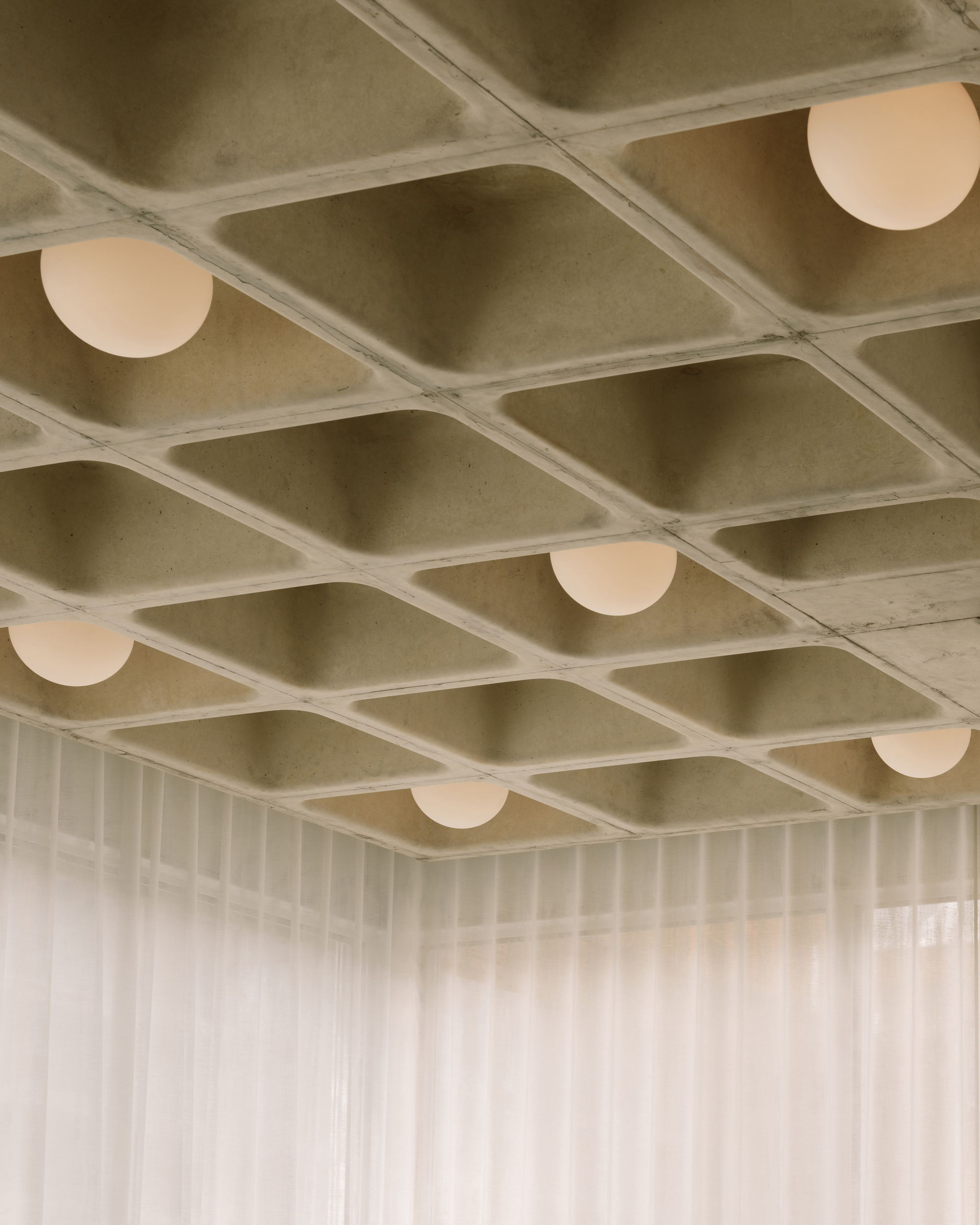 Upward view of waffle slab concrete ceiling with recessed spherical pendant lights and translucent curtain below