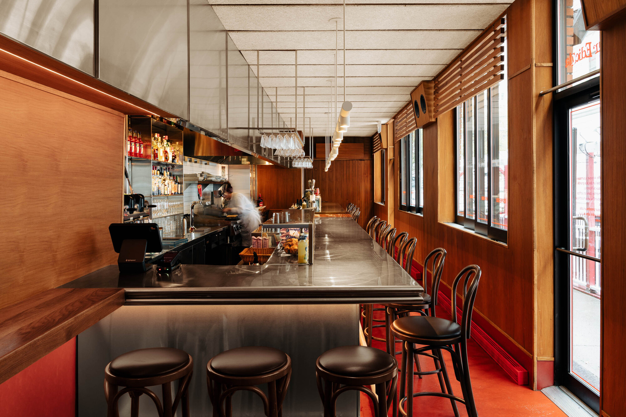 Long stainless steel bar with bentwood stools and red flooring beneath a suspended ceiling grid