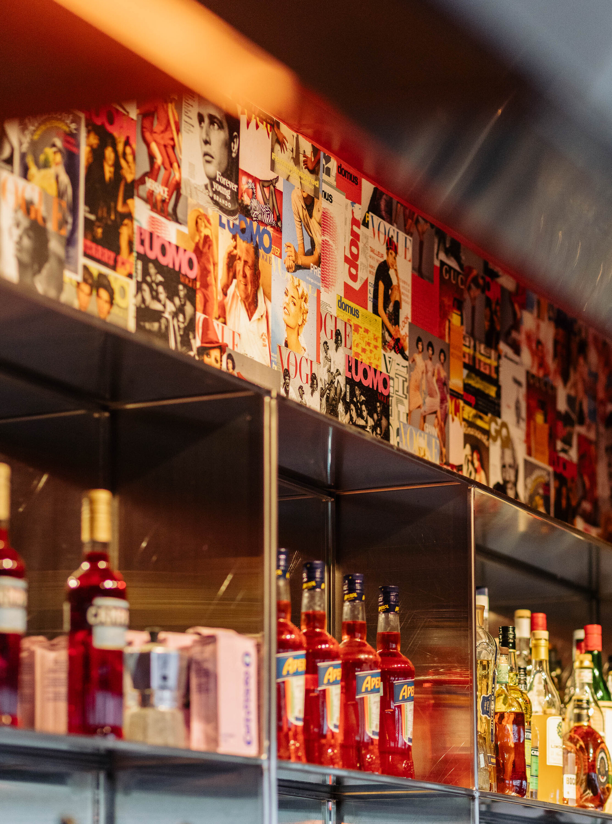 Glass shelving with bottles below a collage band of magazine covers under warm lighting