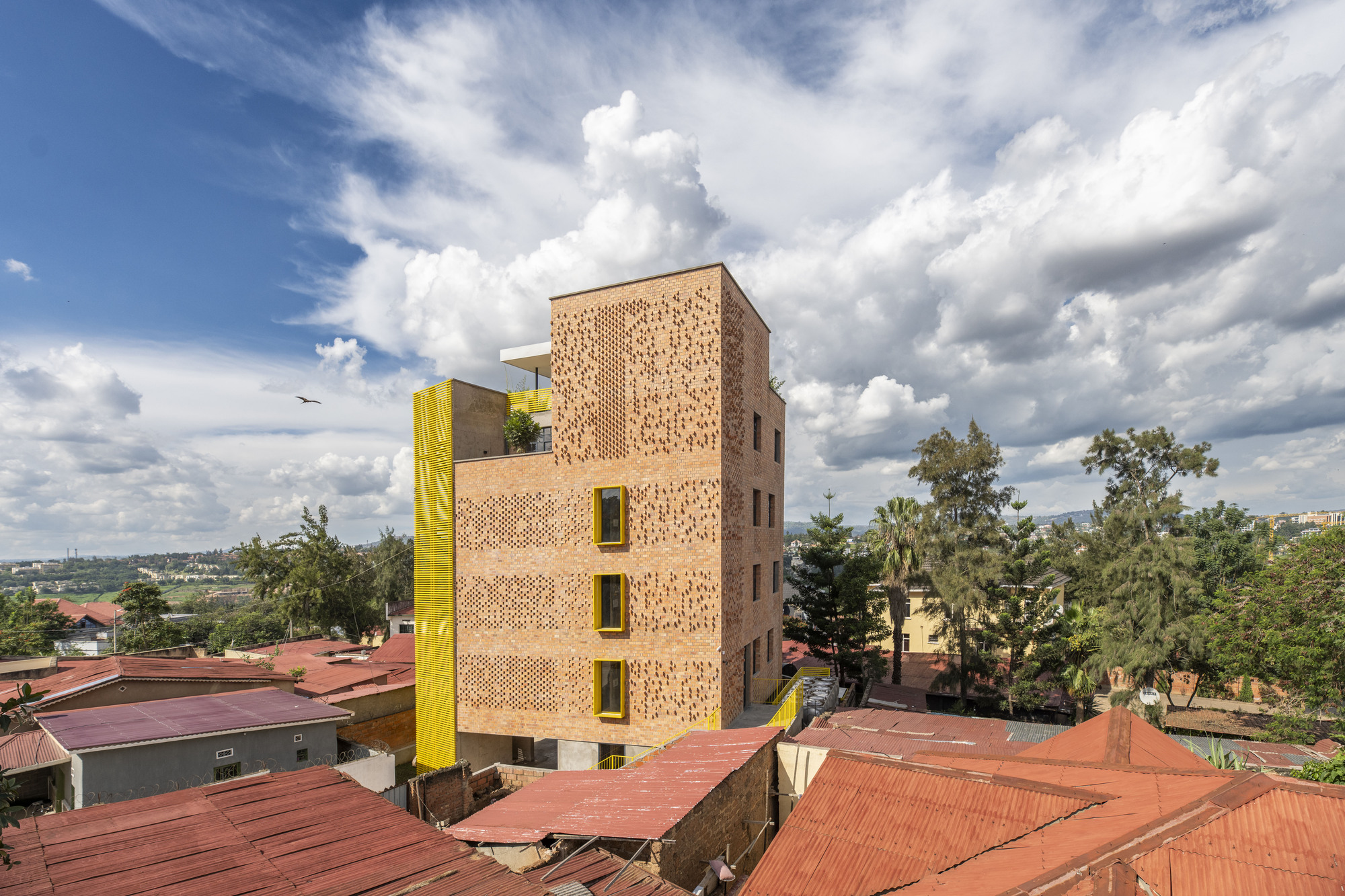 Five-storey brick tower with perforated facade and yellow service column rising above red corrugated roofs