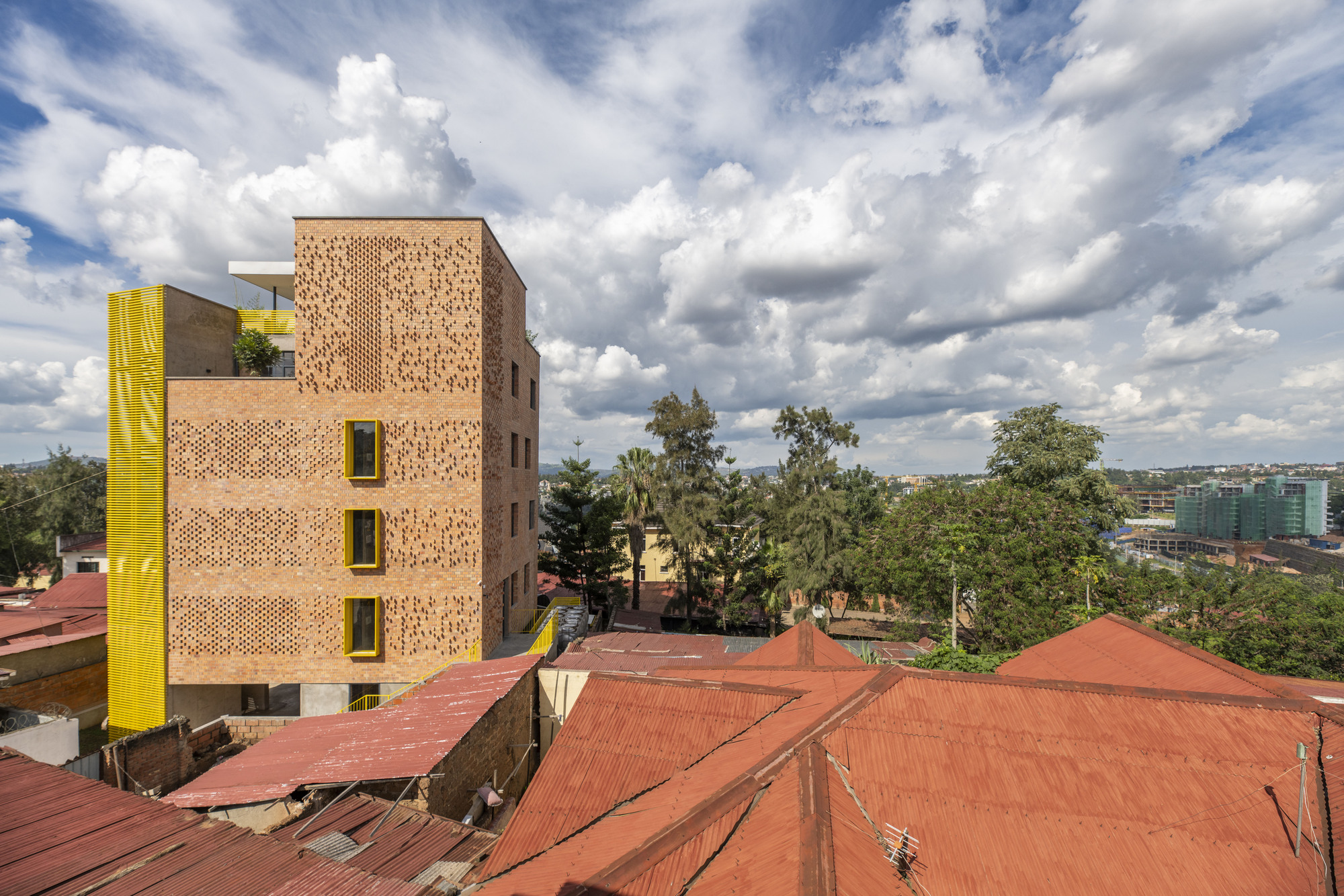 Perforated brick facade with yellow window frames set among trees and low-rise red-roofed buildings