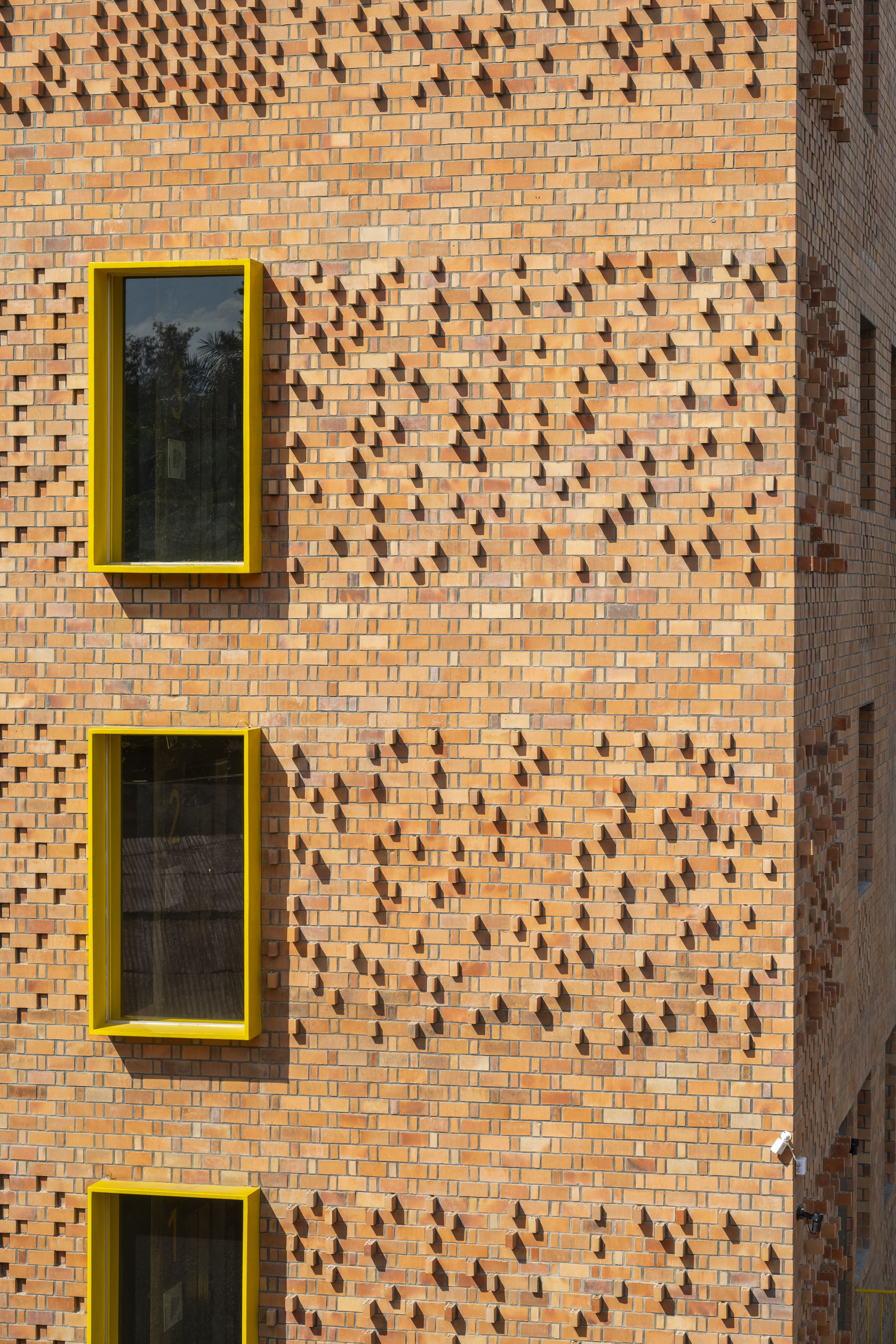 Patterned brickwork facade with protruding units and three yellow-framed windows stacked vertically