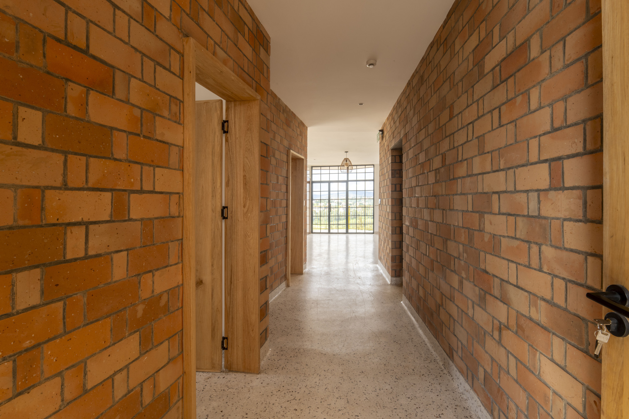 Corridor with exposed brick walls, timber doorframes and terrazzo flooring leading to glazed end wall