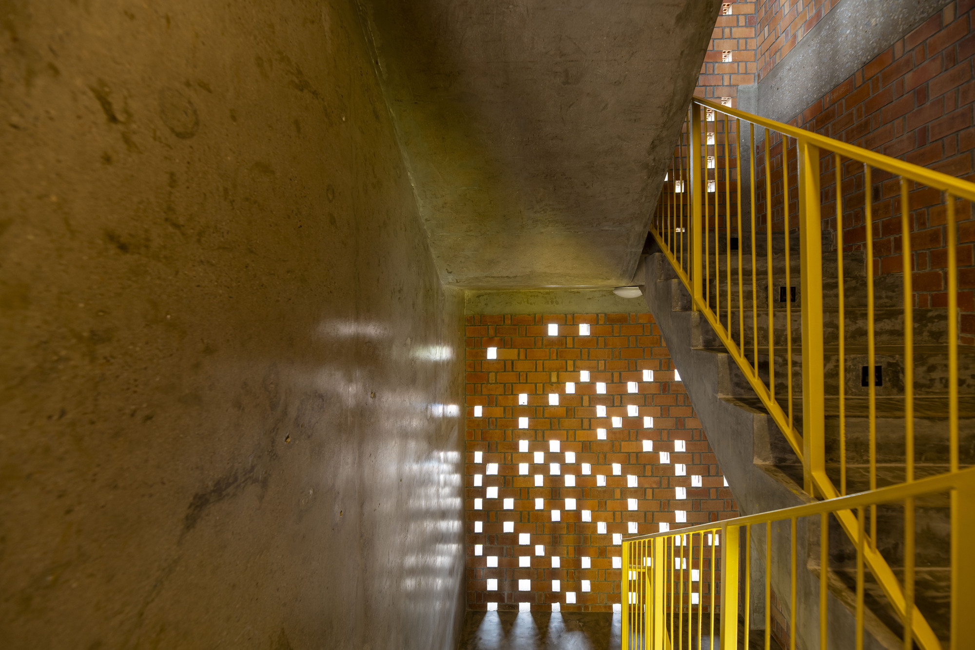 Stairwell with yellow metal railing, concrete walls and perforated brick screen casting light patterns