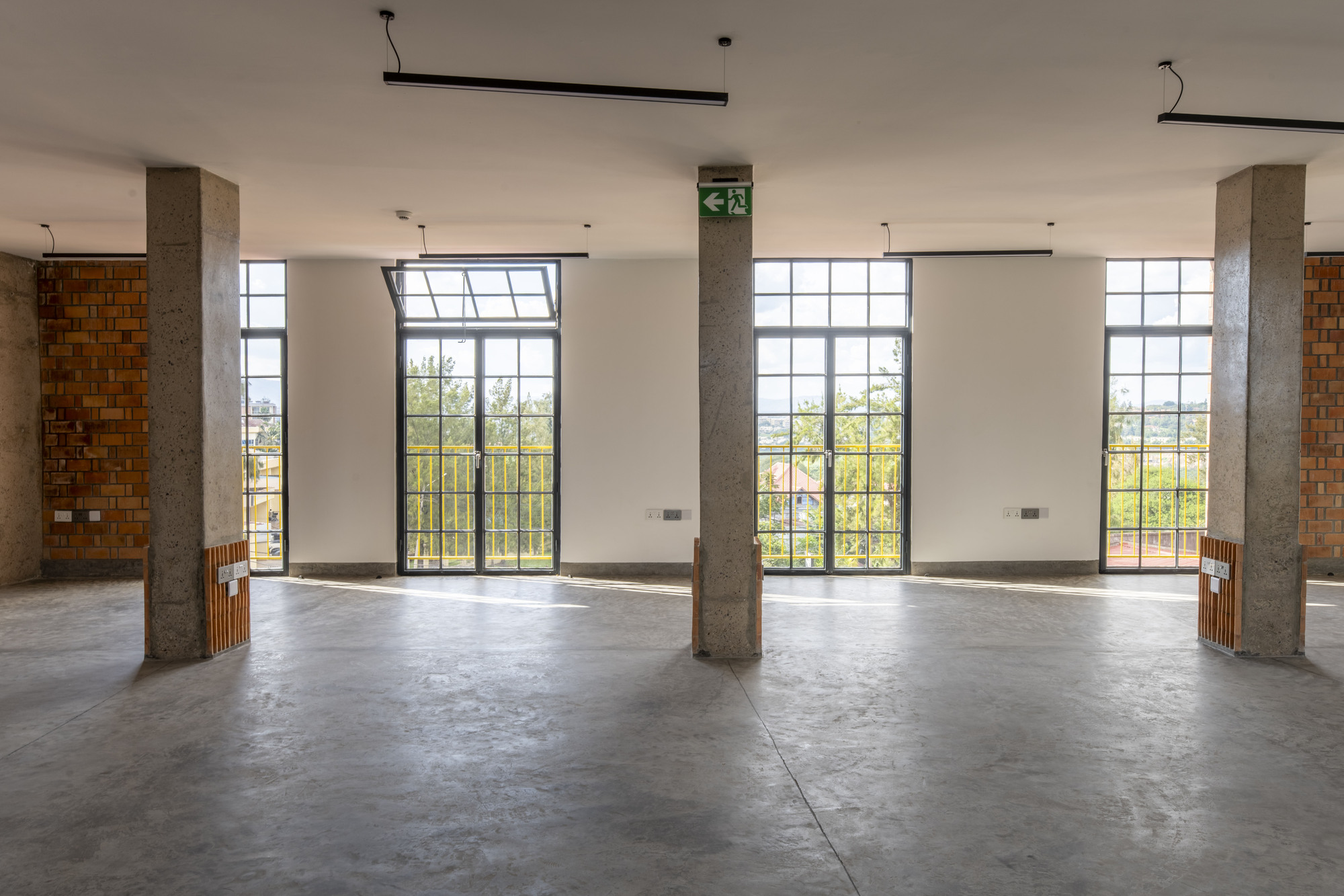 Interior space with three tall gridded windows, concrete columns and timber-wrapped cylindrical elements on concrete floor