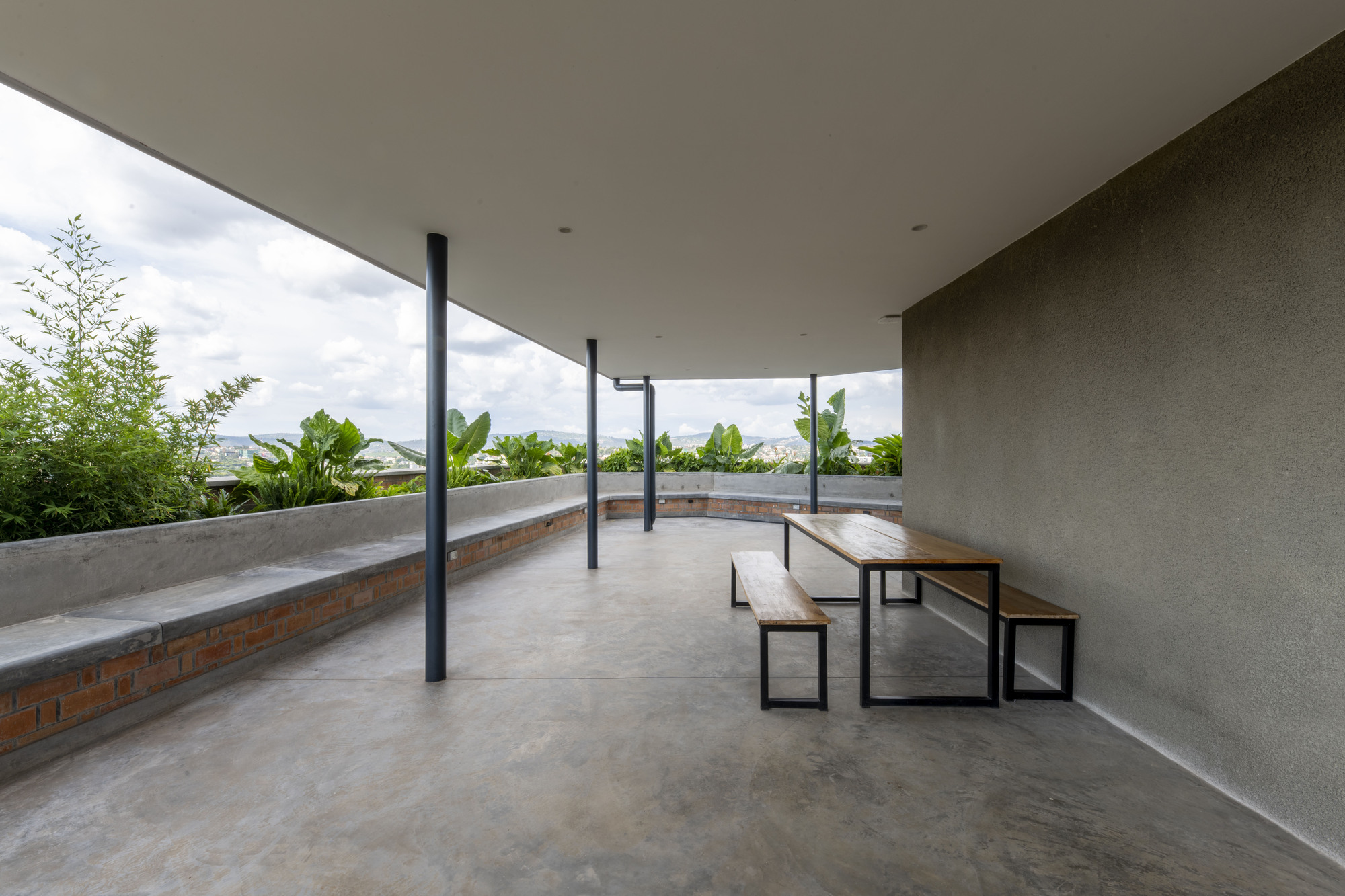 Covered rooftop terrace with curved concrete bench seating, timber picnic table and planted beds under cloudy sky