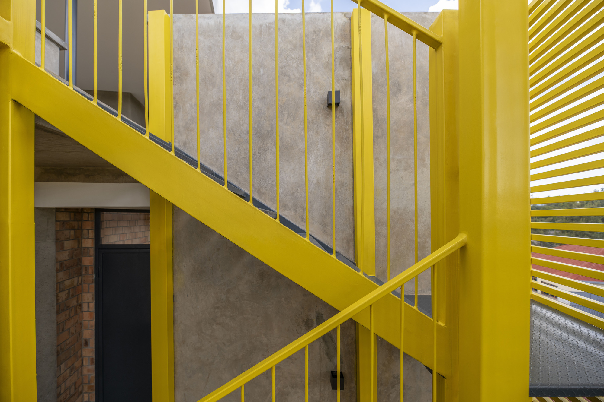 Yellow steel stair with vertical bar railings ascending past concrete and brick walls