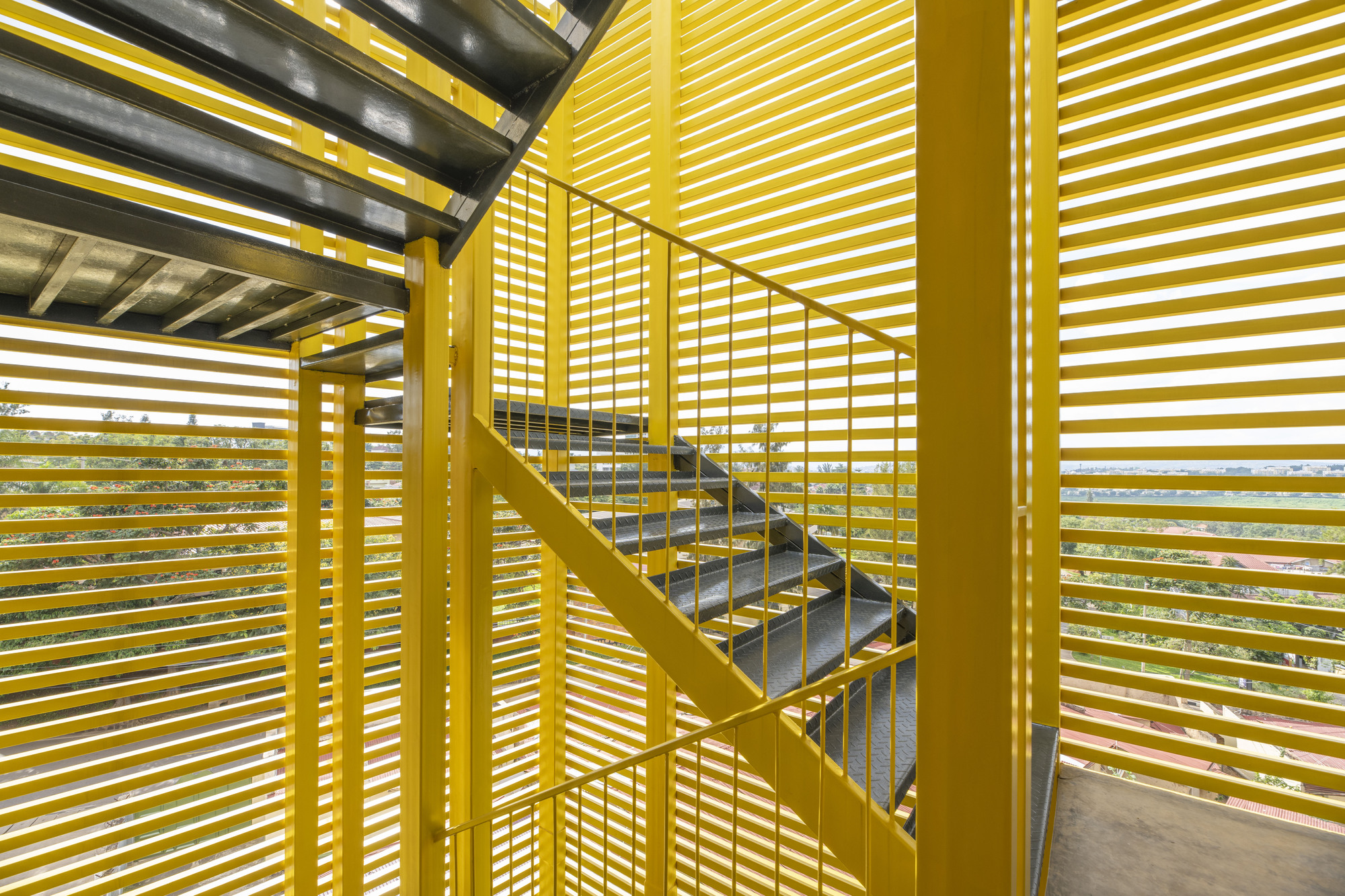 Exterior view through yellow horizontal louvers showing the steel staircase and dappled sunlight