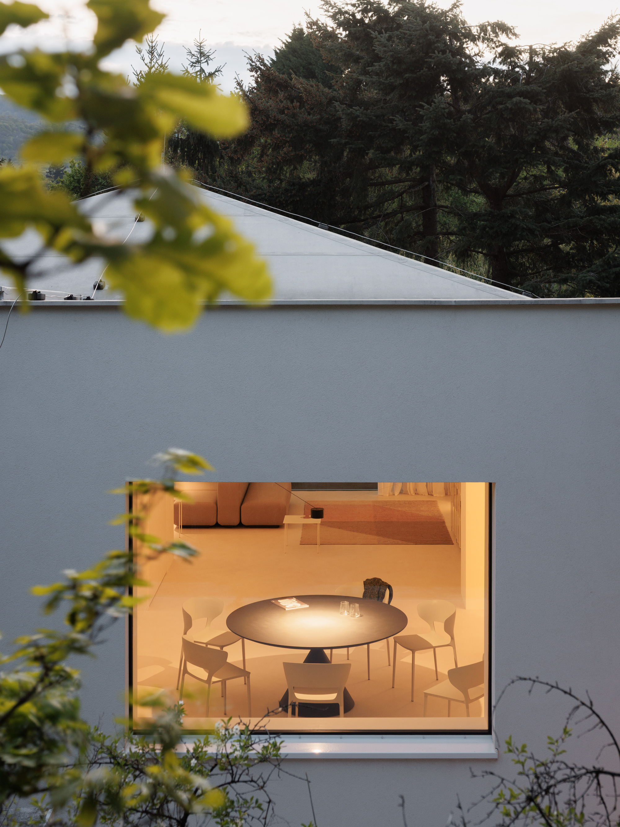Window opening illuminating a dining table at dusk, framed by foreground foliage and roofline